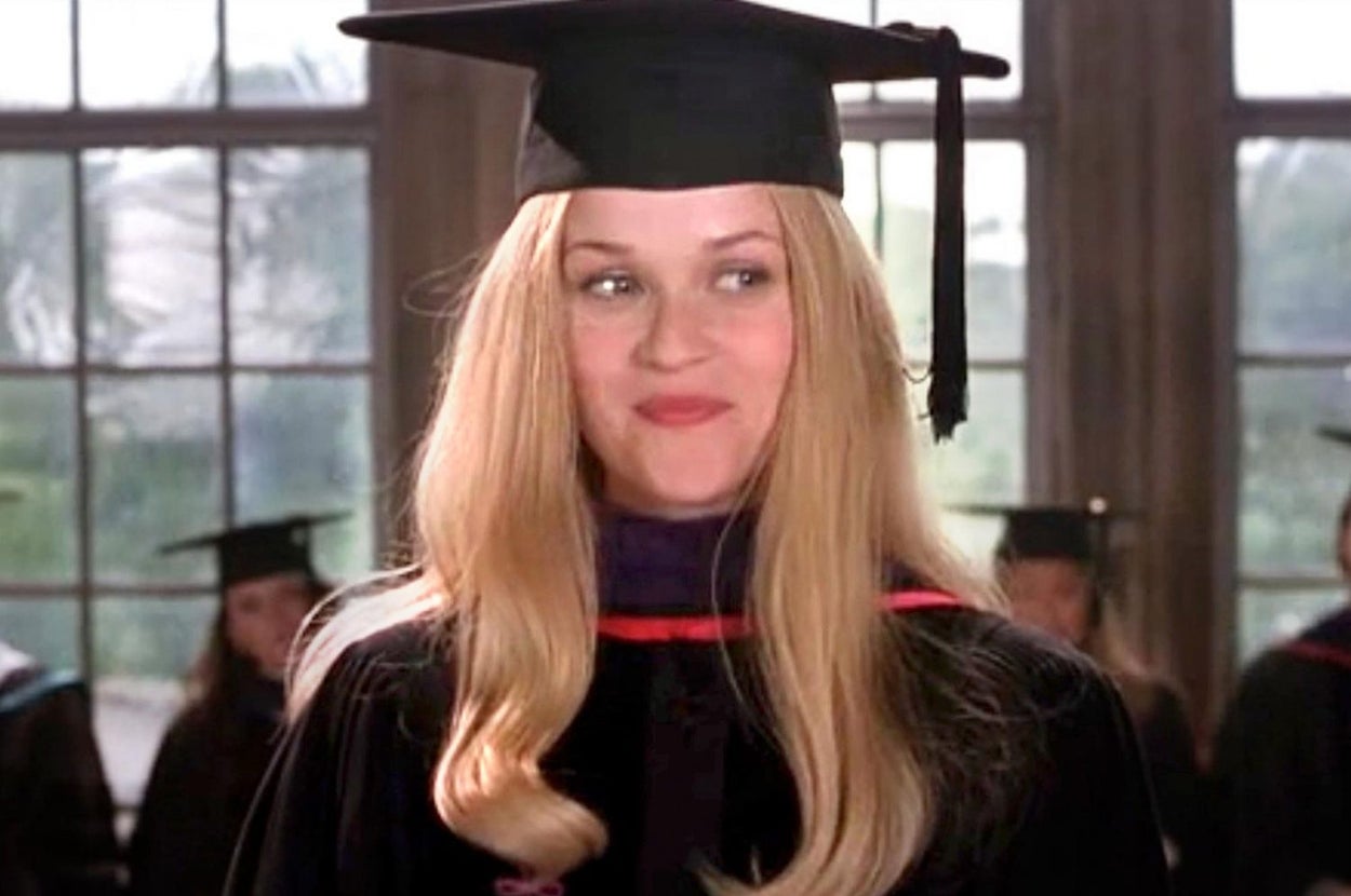 Person in graduation cap and gown smiling indoors, with others in similar attire blurred in the background. Scene from a movie