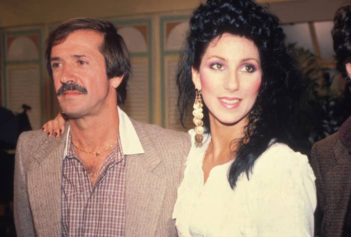 Two people are posing together; one is wearing a blazer and the other a white outfit with large earrings