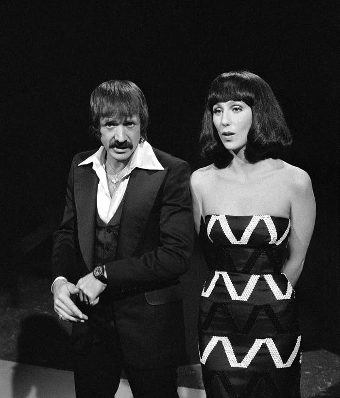 Two people stand together. The person on the left wears a suit and open-collared shirt. The person on the right wears a strapless zigzag-patterned dress