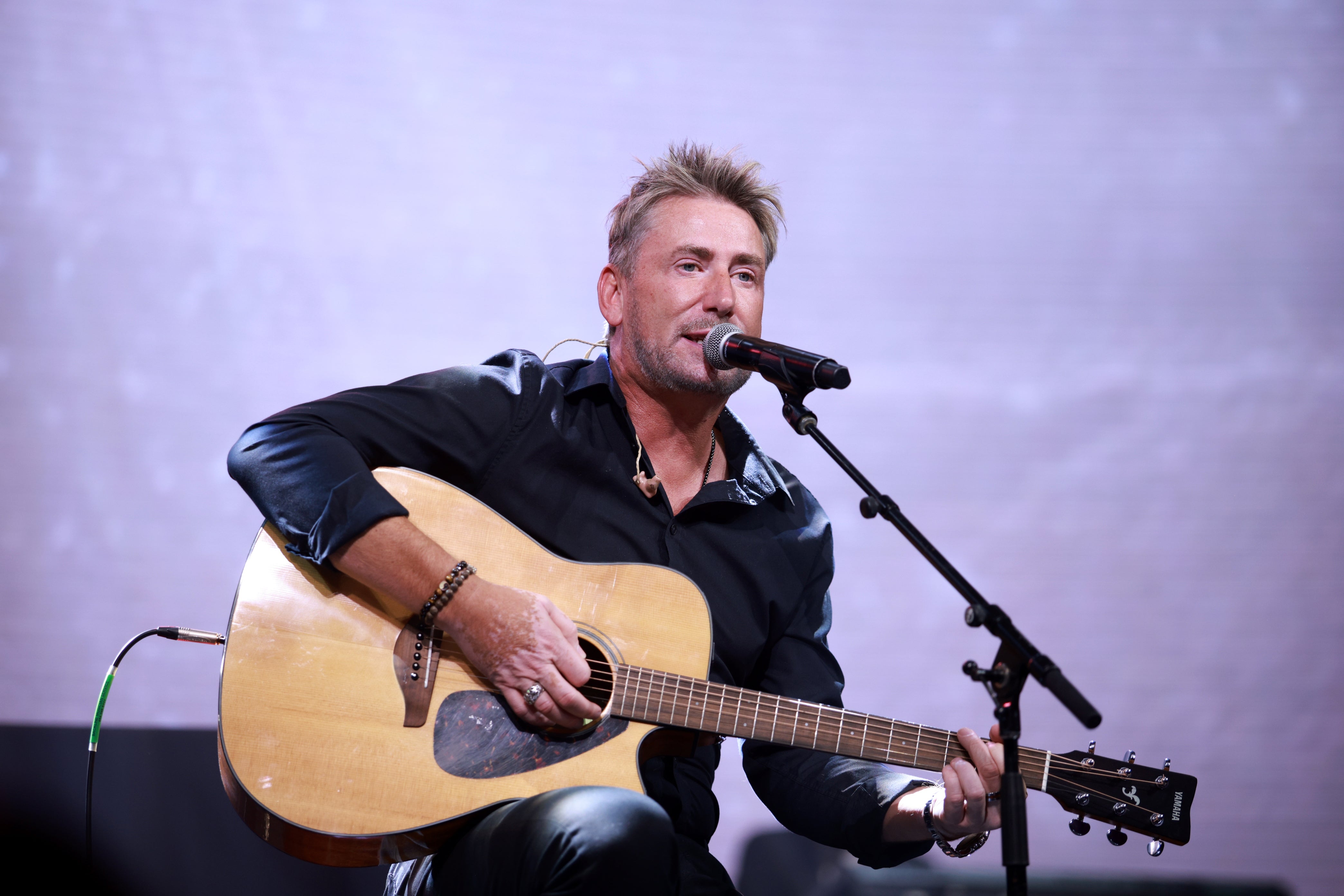 A man performs on stage, playing an acoustic guitar and singing into a microphone, wearing a casual dark shirt