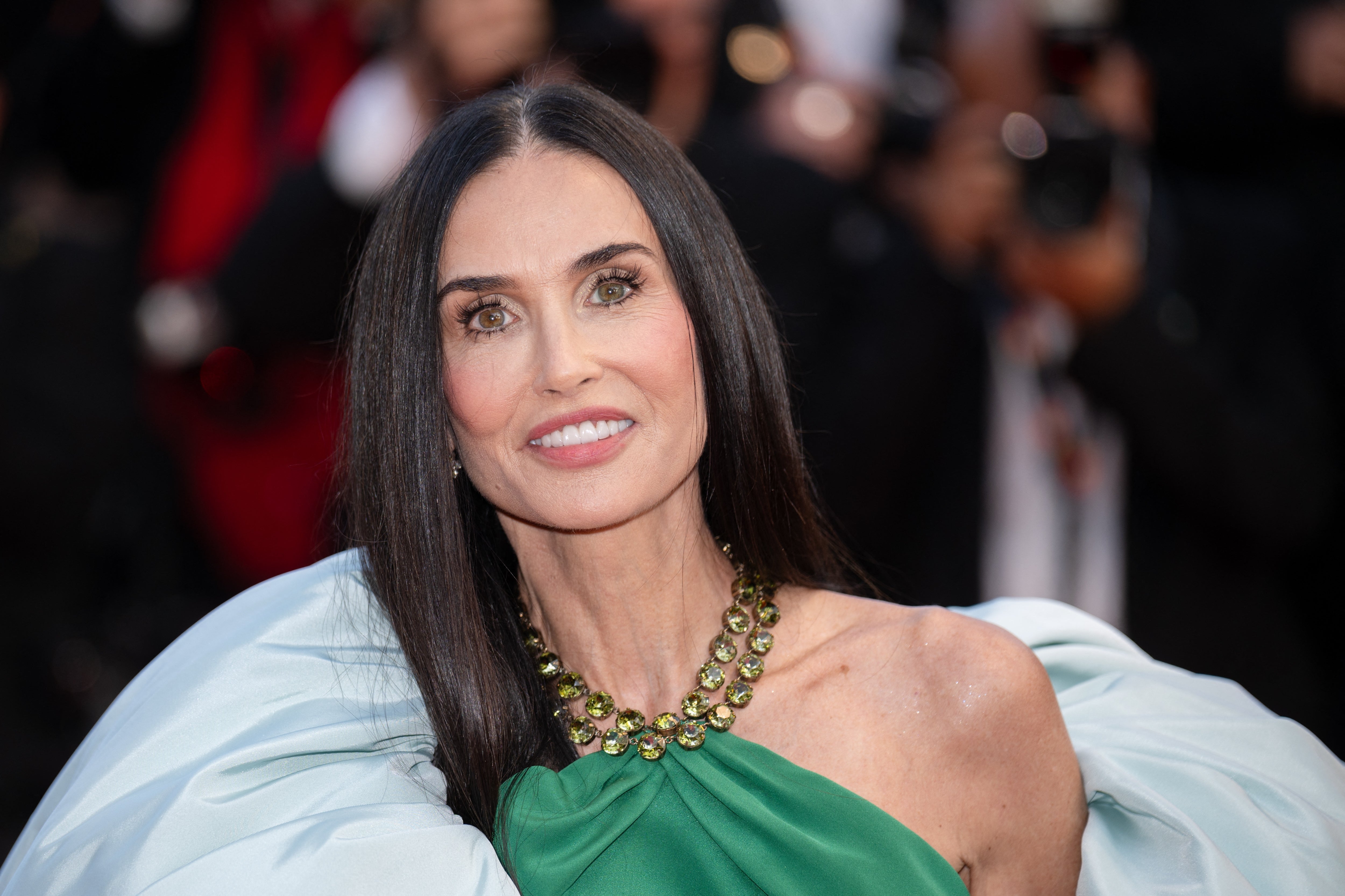 A woman in a strapless gown with a statement necklace poses on a red carpet with photographers in the background