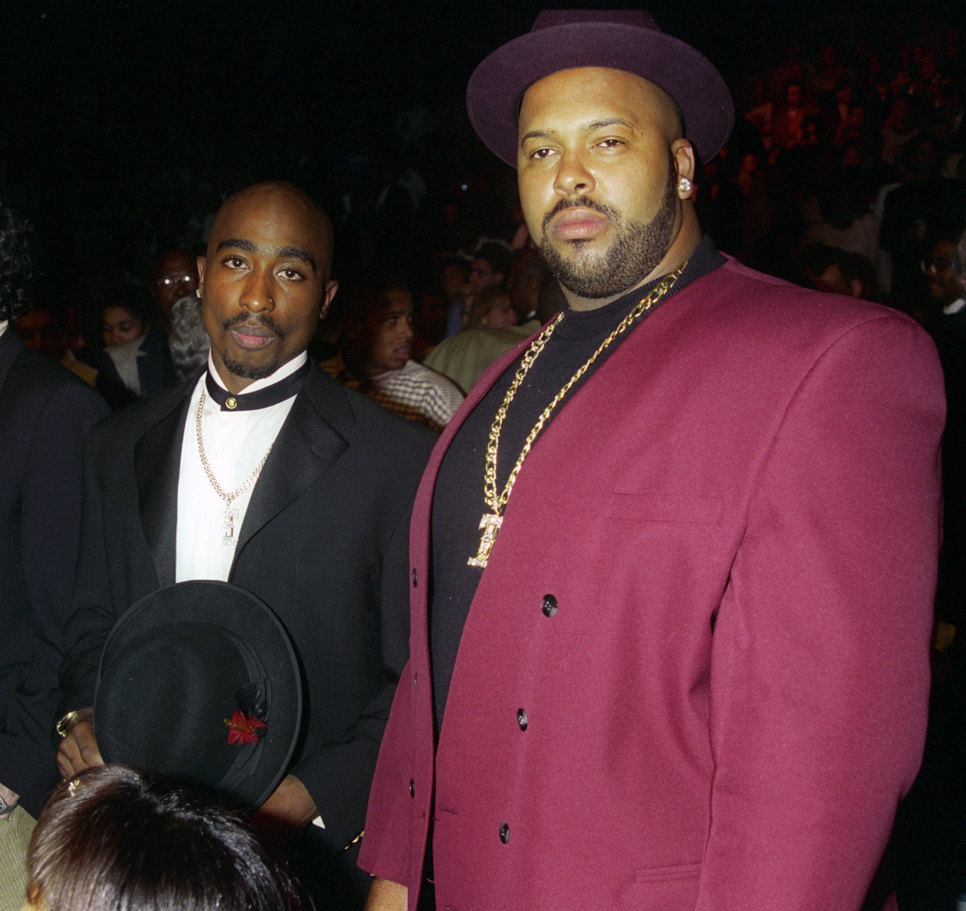 Two individuals in stylish suits and hats pose together at an event, one with a chain necklace