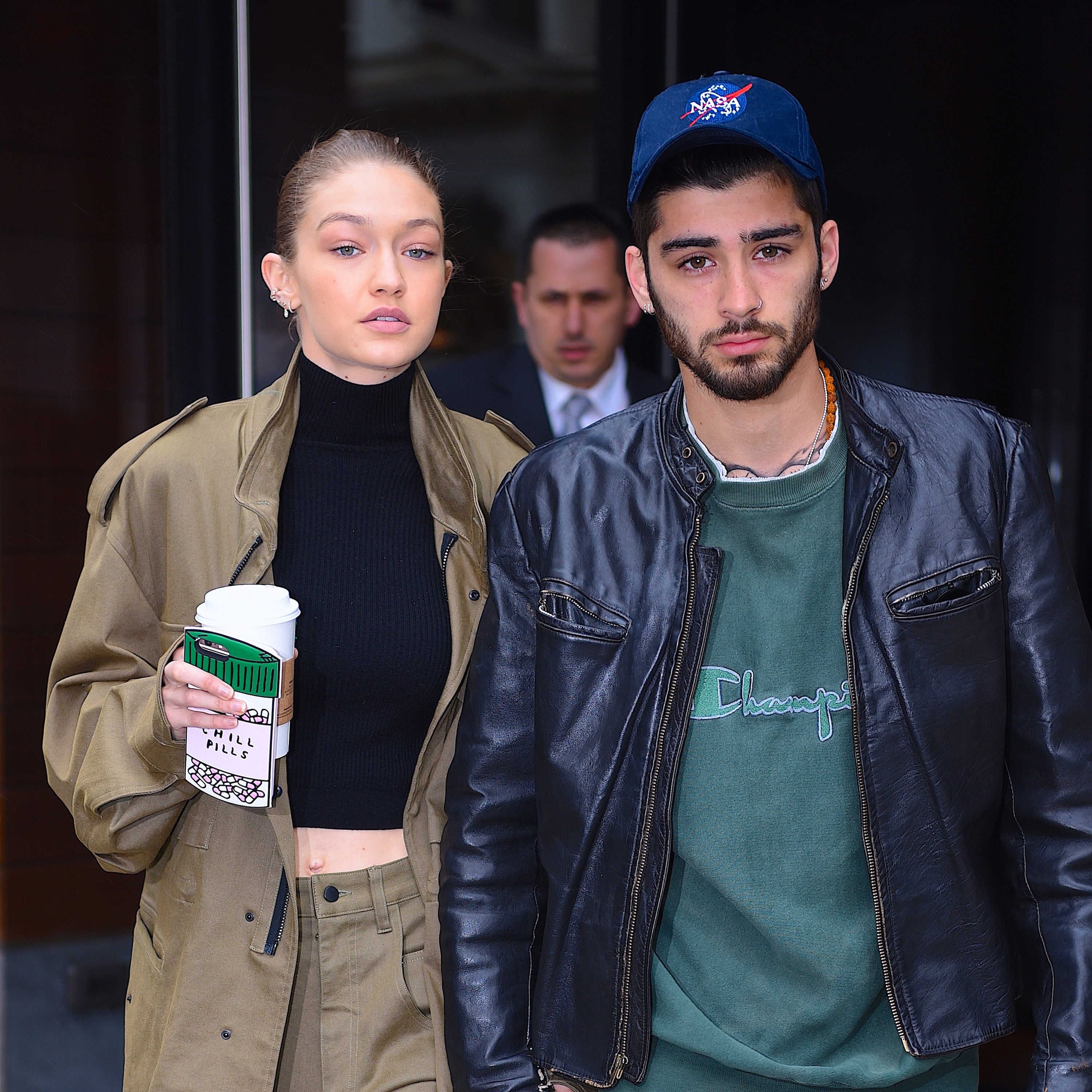 Two people walking outdoors; one wears a trench coat and holds a coffee cup, the other wears a leather jacket and a cap