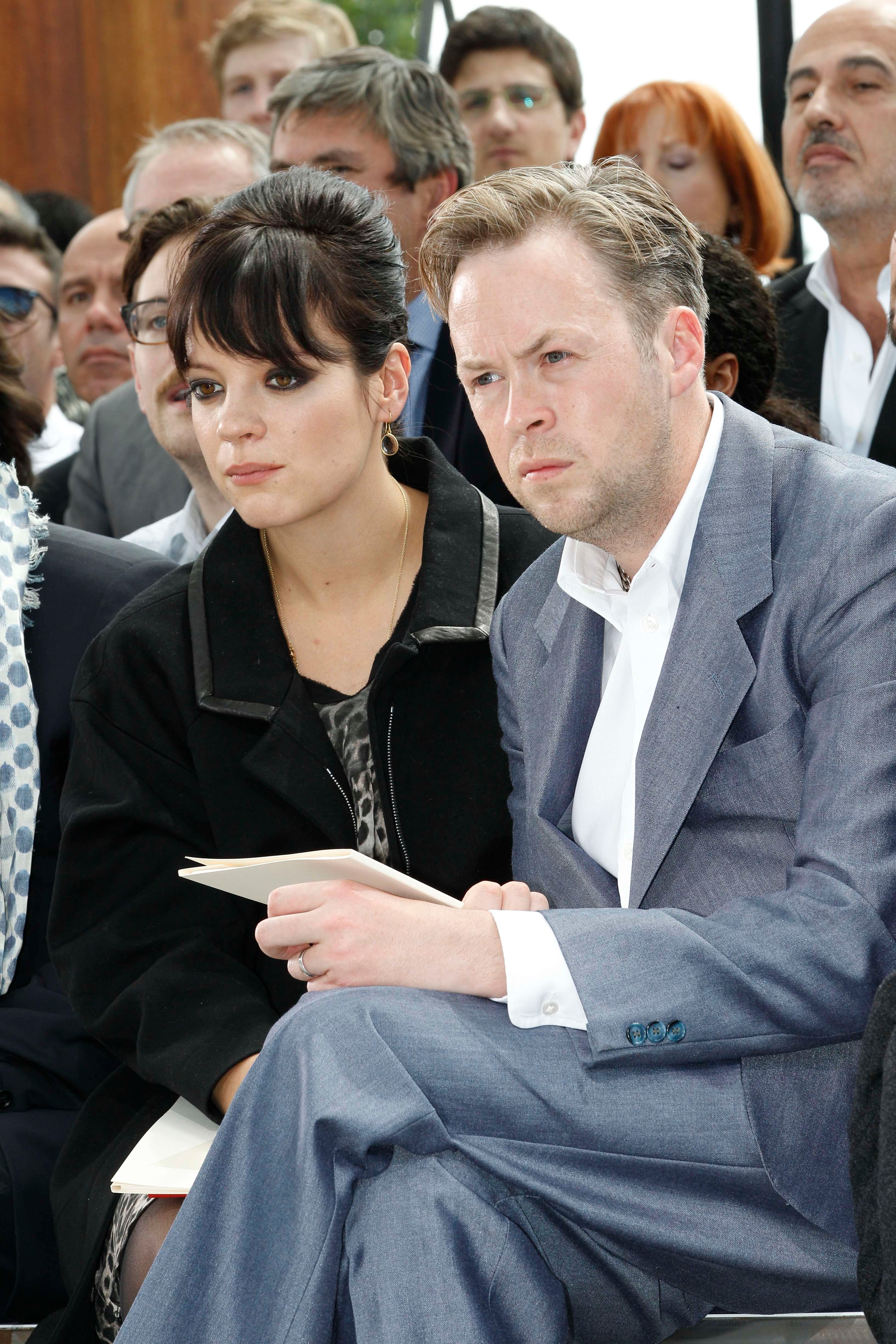 Two individuals seated together, dressed formally. One has dark hair and wears a black jacket; the other has lighter hair and wears a gray suit