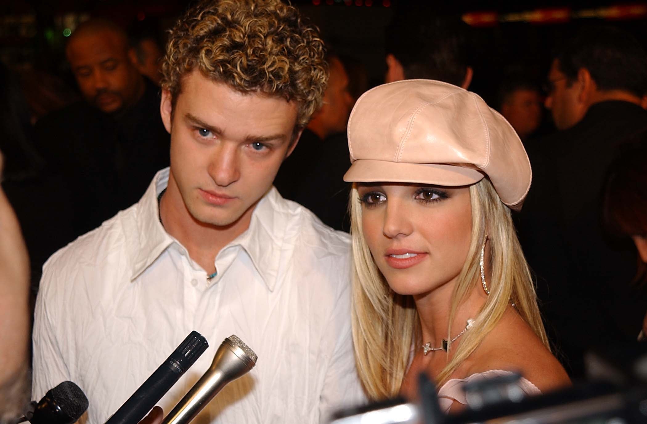 Two celebrities pose together, one wearing a white shirt and the other in a beret with a strapless top, surrounded by microphones