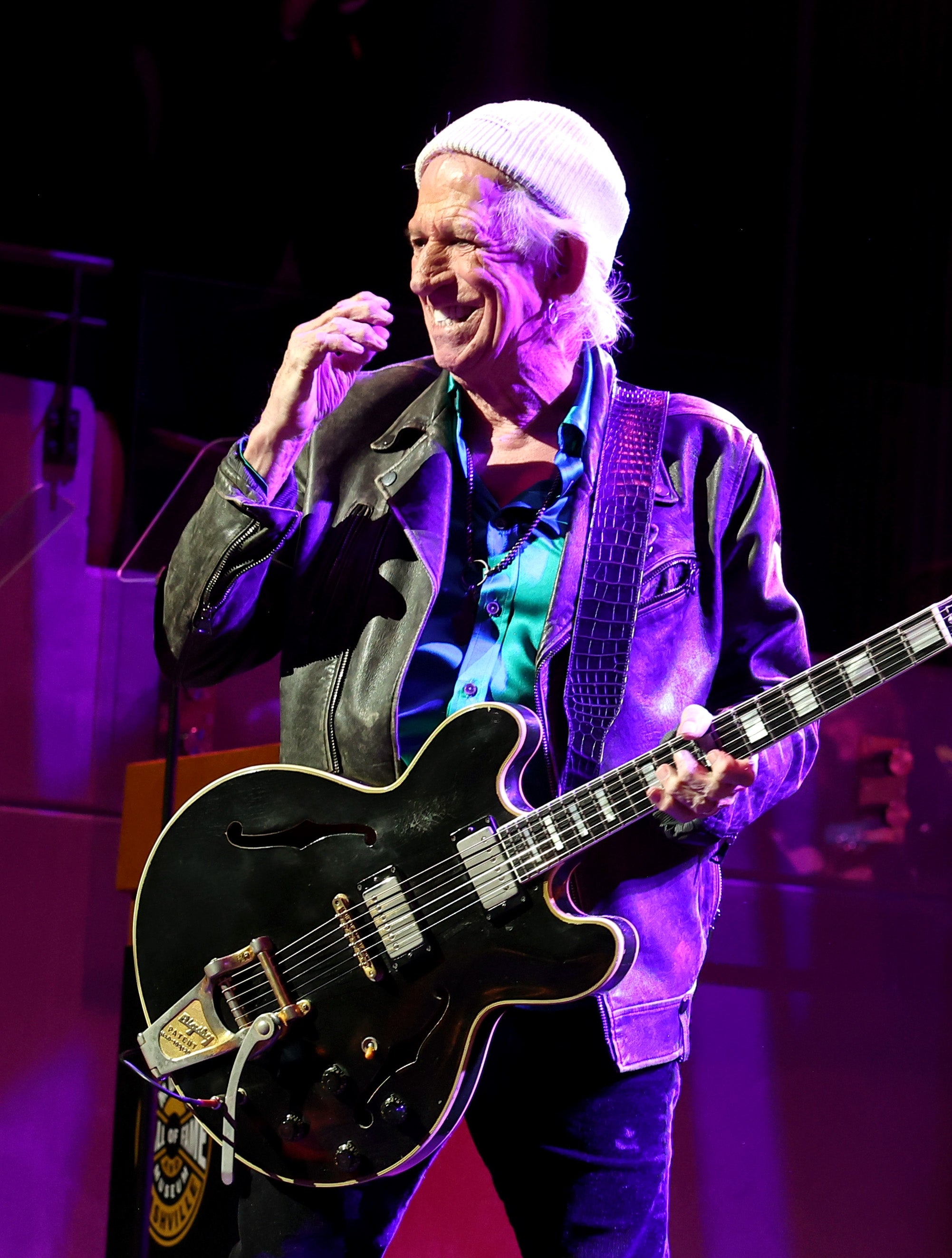 Guitarist wearing a leather jacket and beanie performs energetically on stage, holding an electric guitar and smiling
