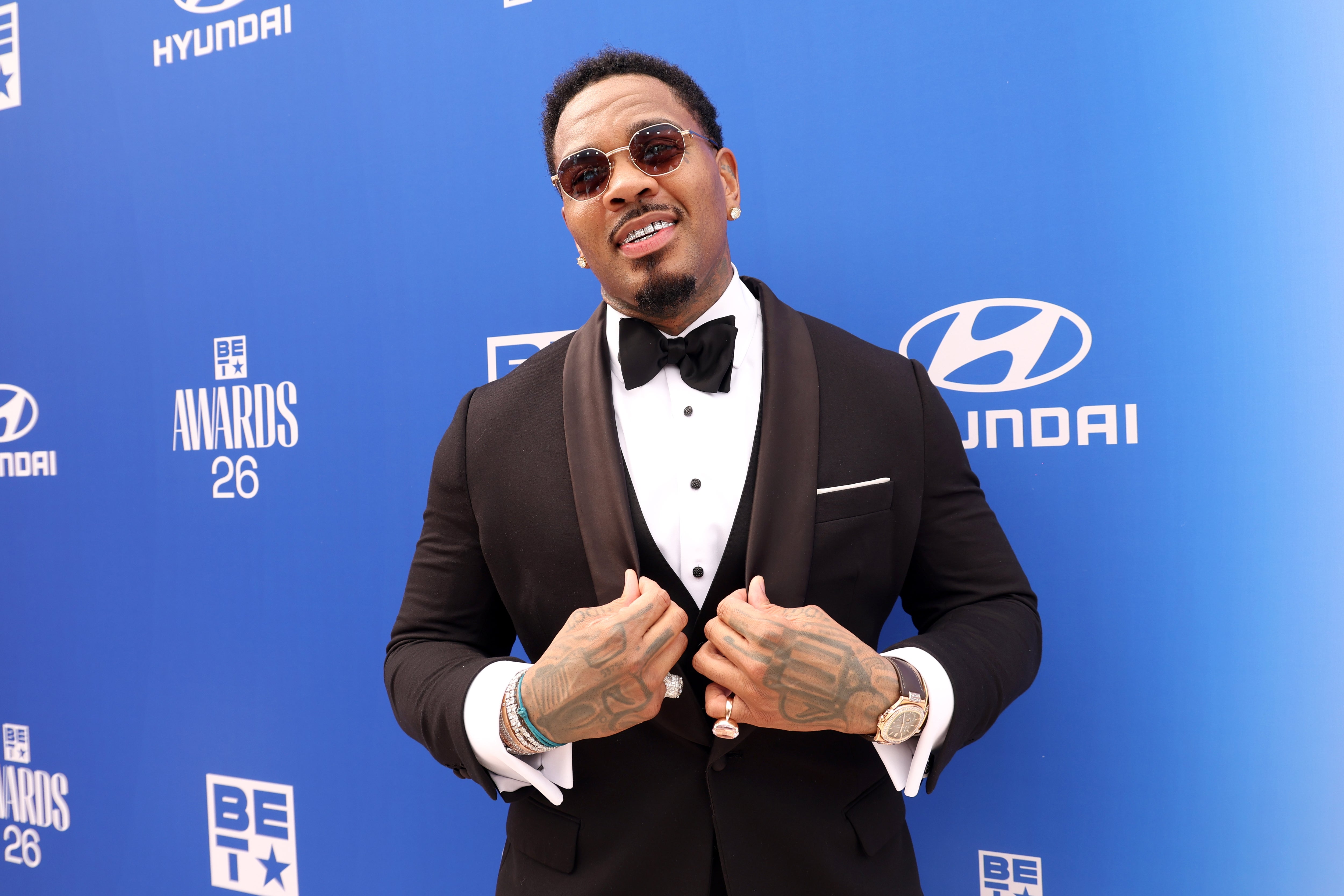 A person in a sharp tuxedo poses confidently on a blue carpet at an awards event, showcasing tattoos and wearing sunglasses