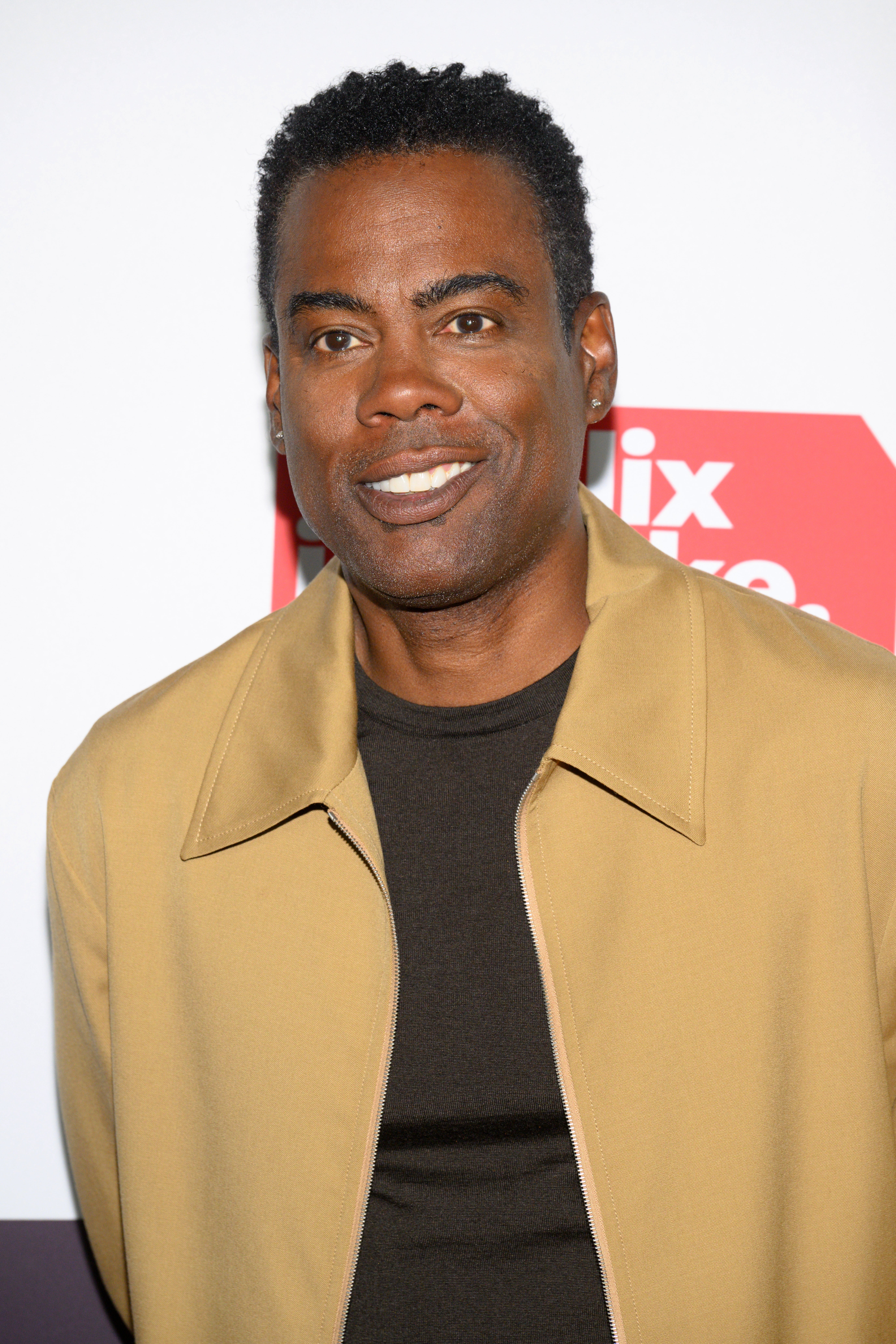 Man in a tan jacket and dark shirt at a media event, smiling at the camera