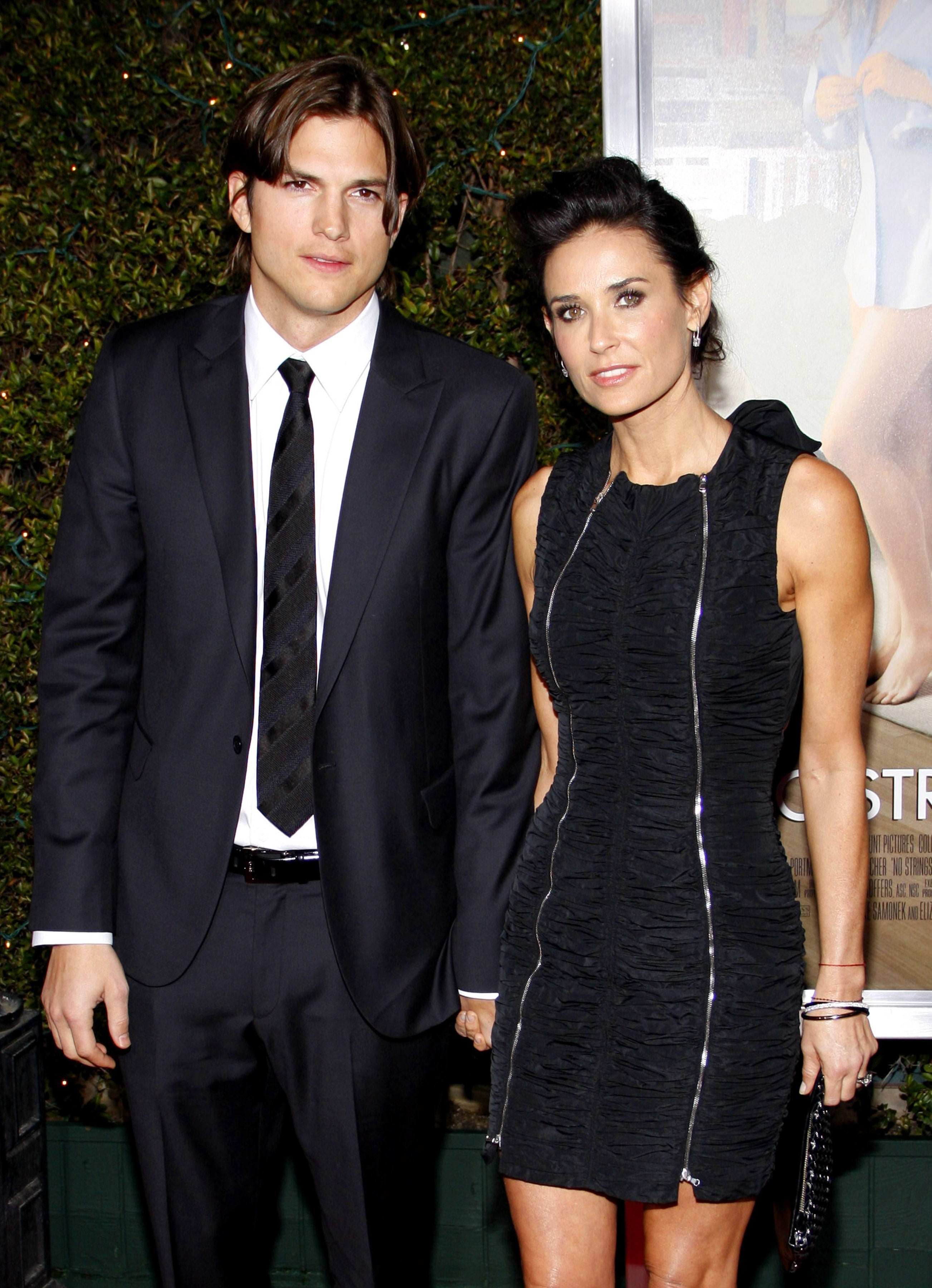 Two people pose together. The man wears a black suit and tie, while the woman is in a sleeveless, textured black dress holding a clutch