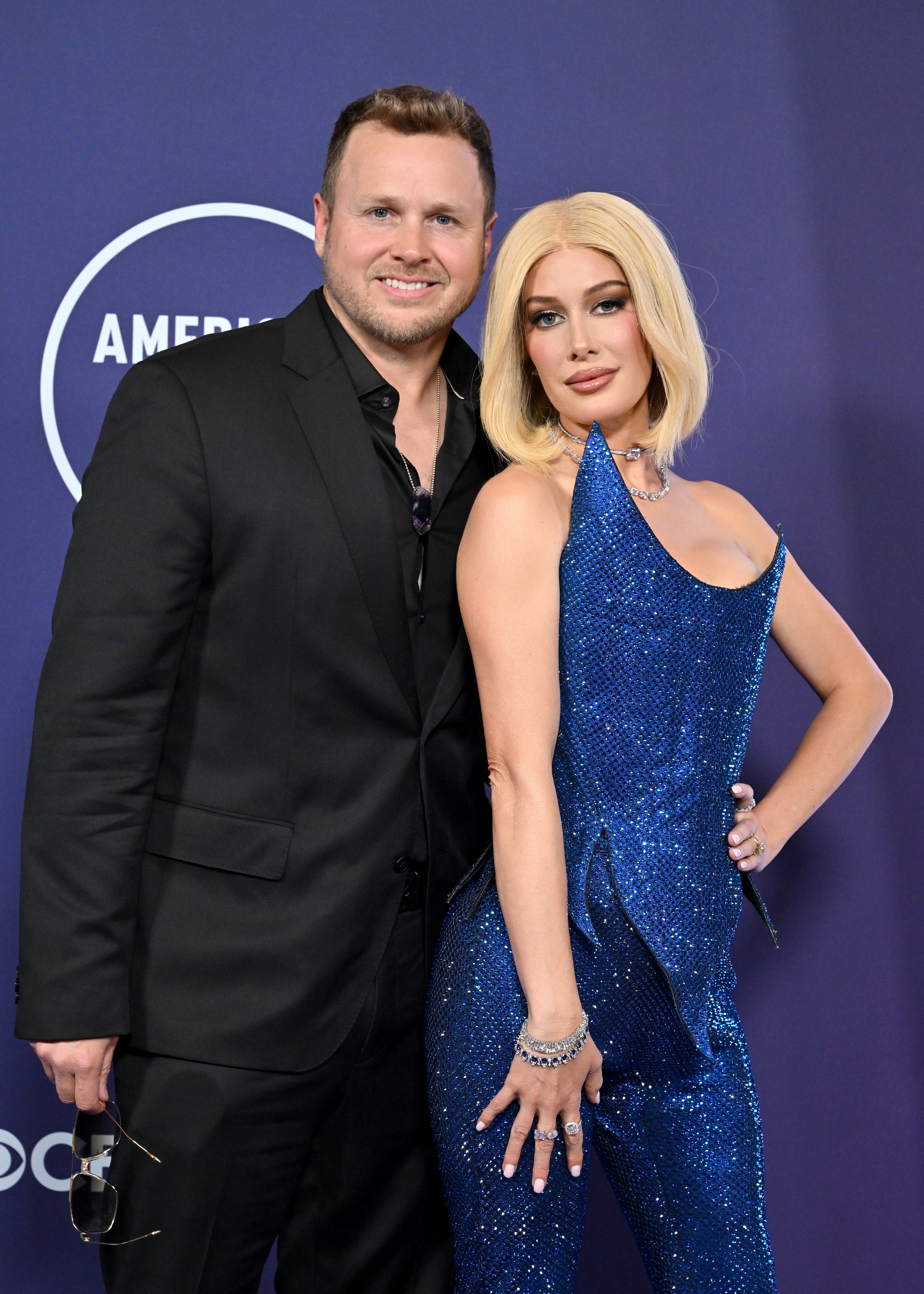Two people pose at an event. The person on the left wears a black suit, and the person on the right wears a sparkly, form-fitting dress