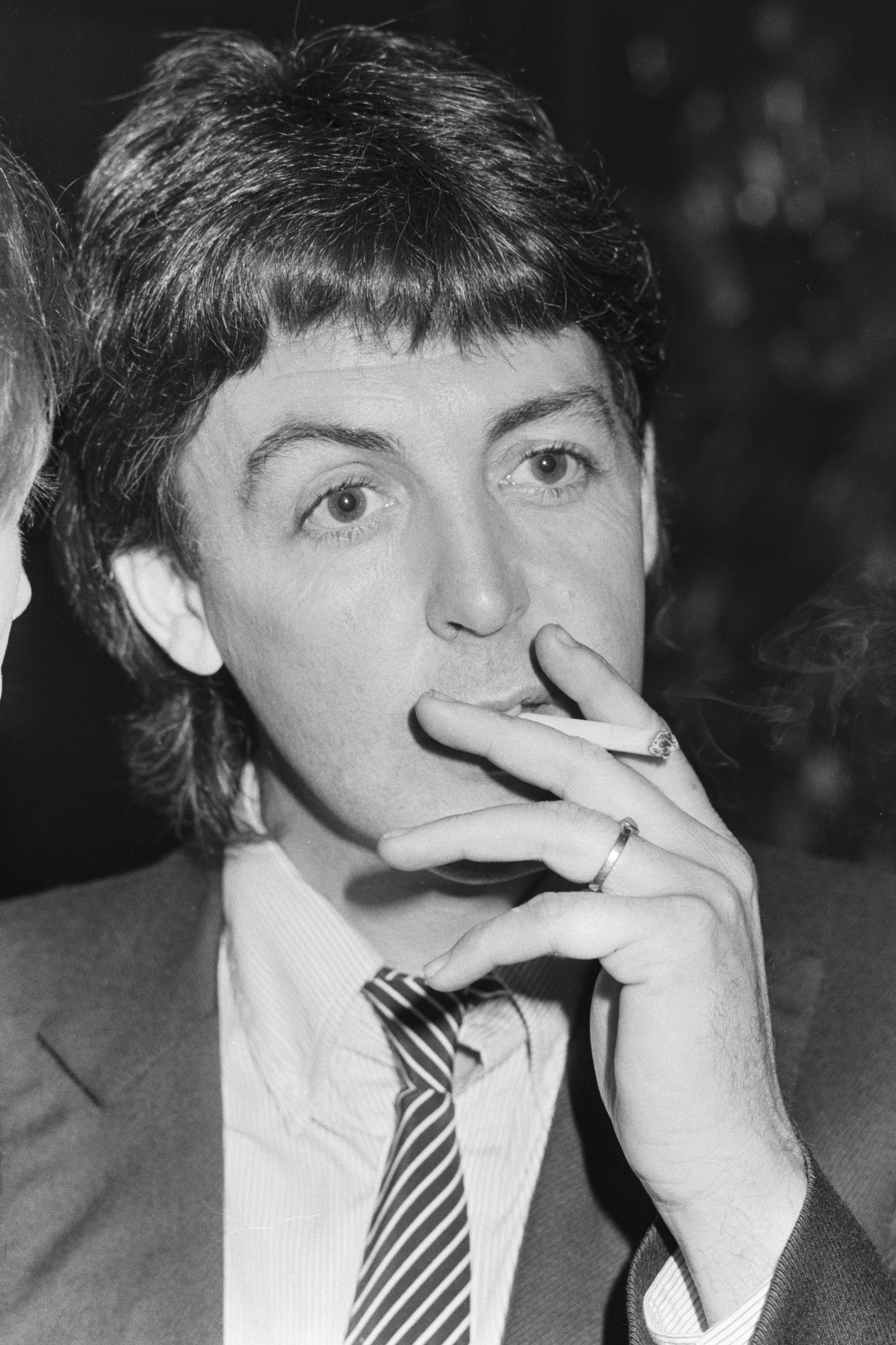 Person in suit with striped tie, holding a cigarette near their mouth, looking thoughtful