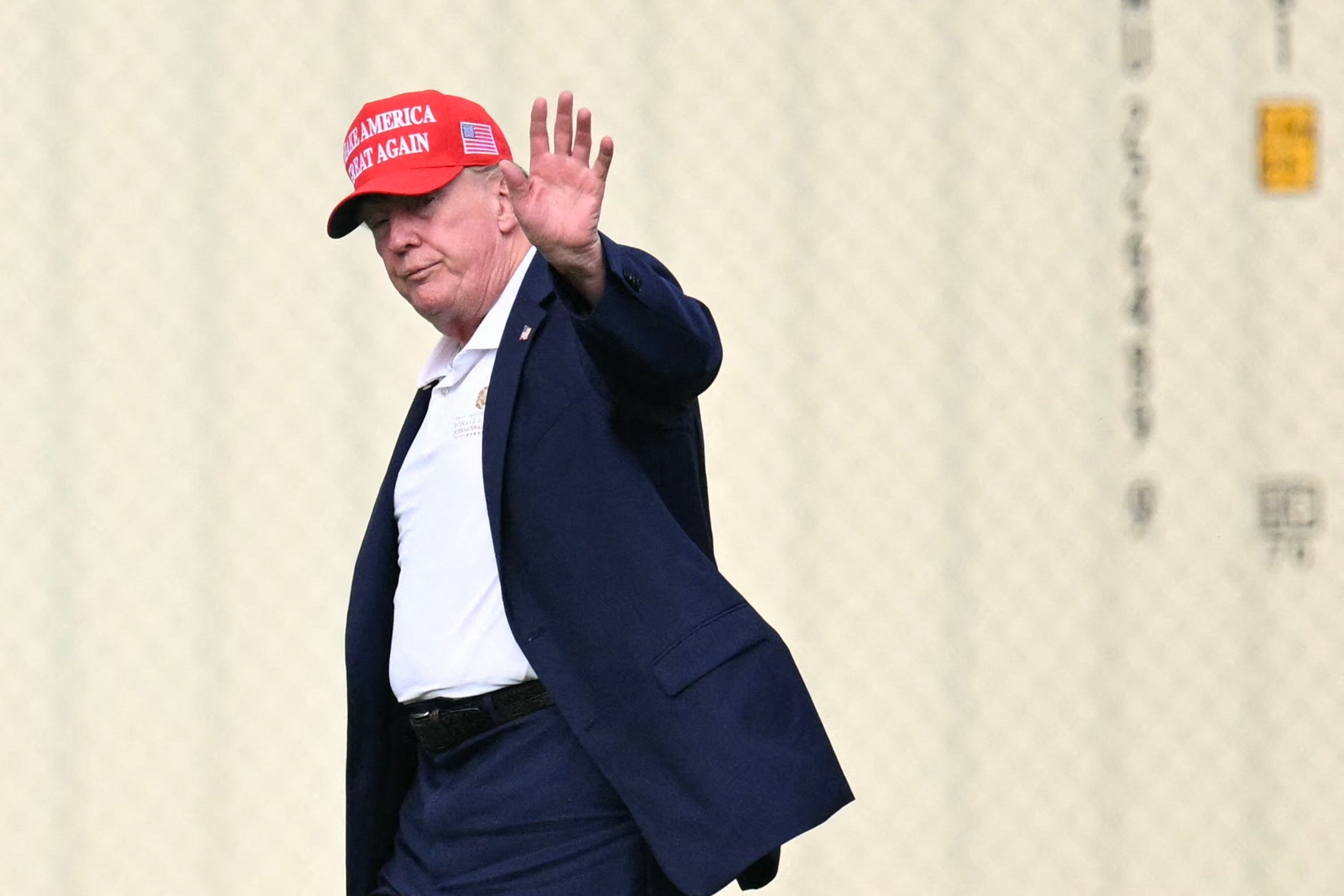 Mann i dress vinker, iført en rød "Make America Great Again" cap, mot en vanlig bakgrunn