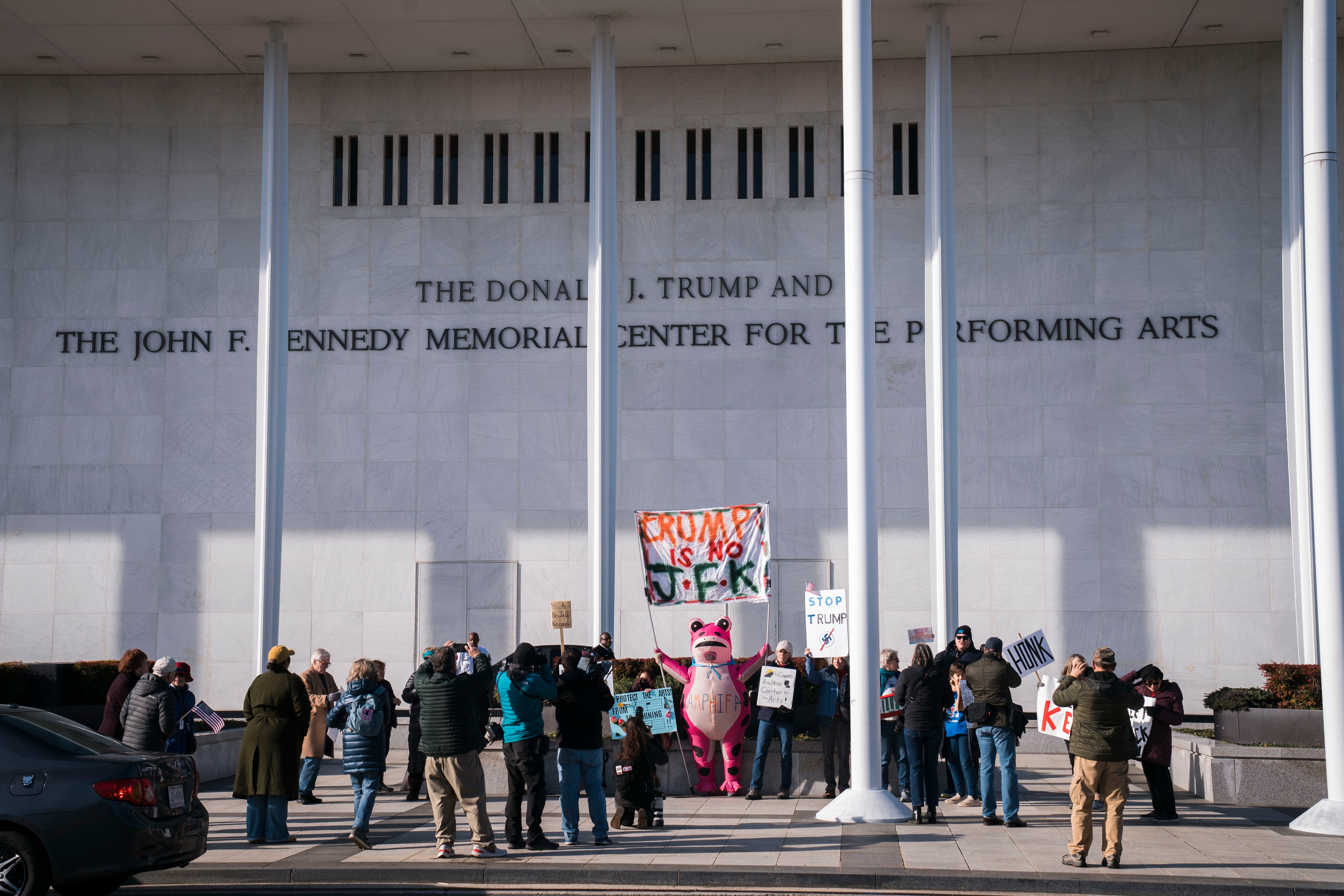 En gruppe demonstranter med skilt, inkludert en med en rosa dinosaur, står utenfor Kennedy Center