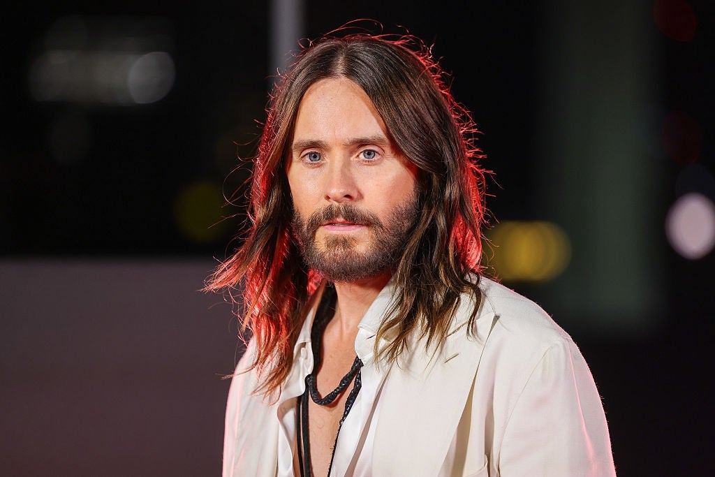 Person with long hair and beard in a white suit and black necklace on a red carpet