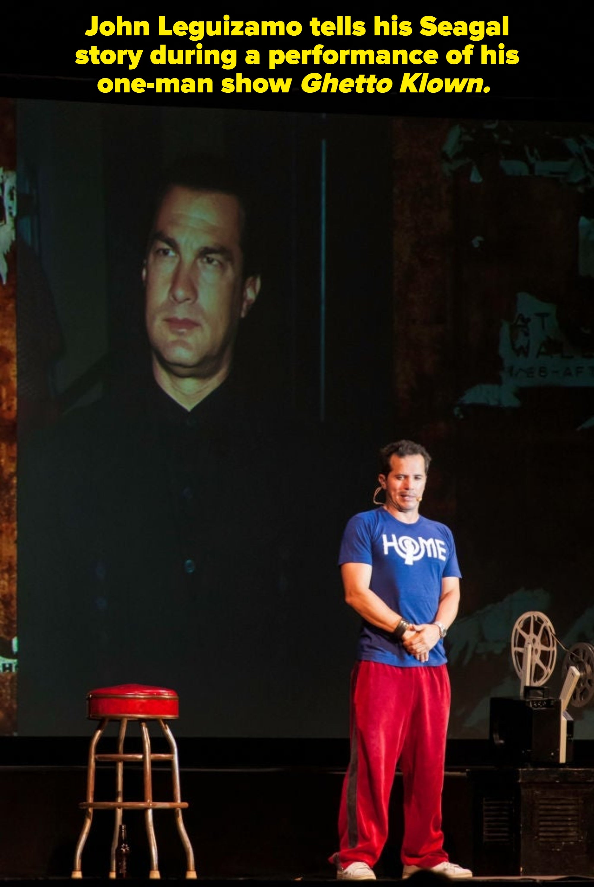 Person on stage wearing a "Home" t-shirt and red pants stands in front of a large projection of another man's portrait