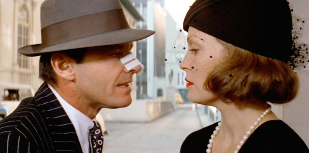 A man in a pinstripe suit and hat faces a woman wearing a hat and pearl necklace, both in a dramatic, vintage style setting