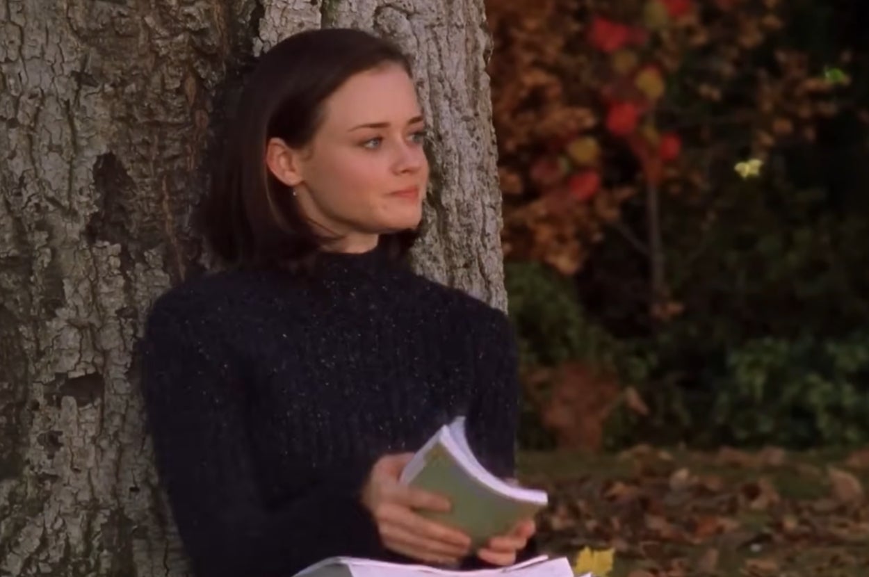 Rory Gilmore from "Gilmore Girls" sitting next to a tree, holding a book, in an outdoor setting with fallen leaves
