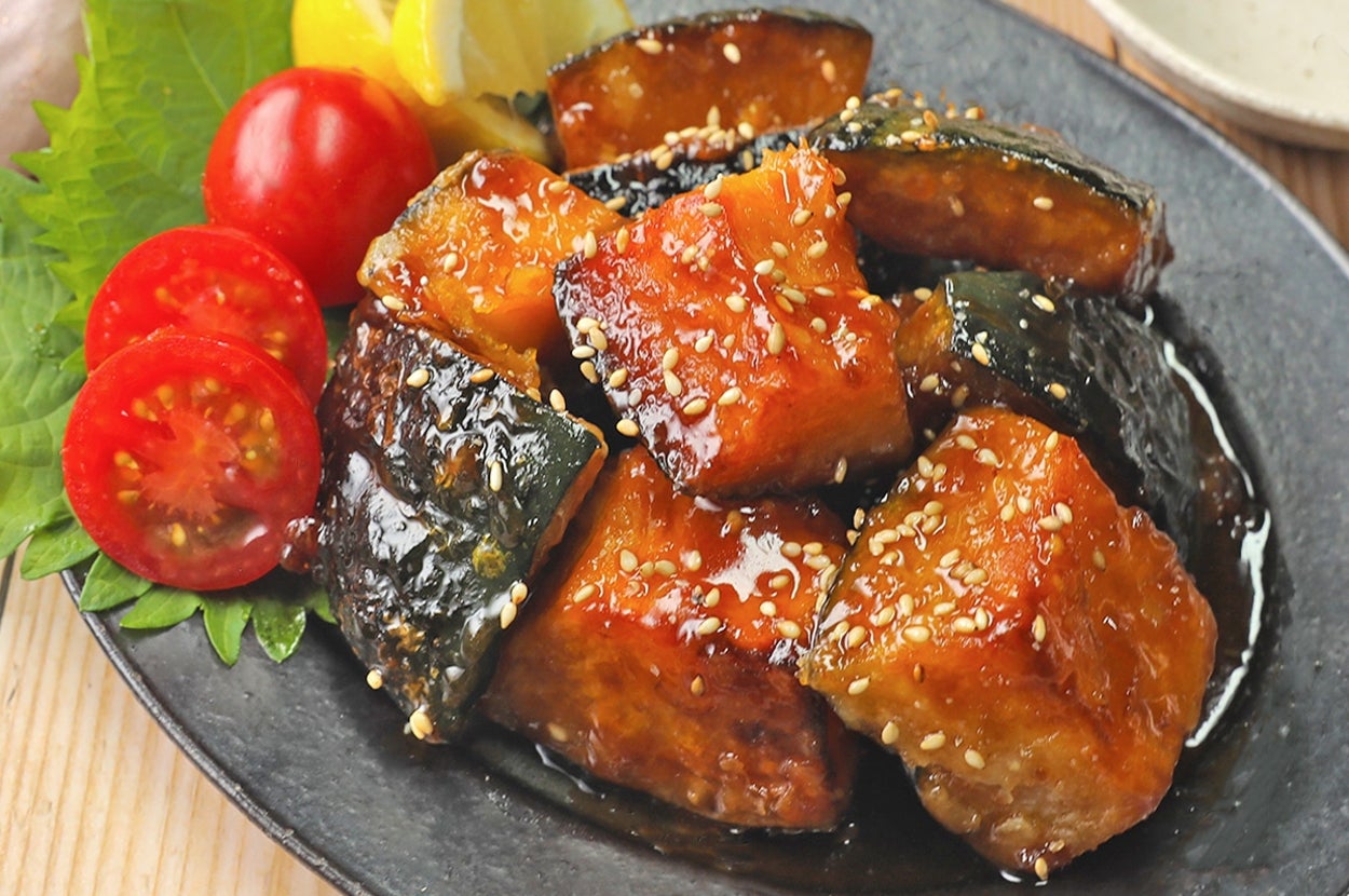 皿に盛られたてりやき味のかぼちゃと豆腐の煮物。ゴマがまぶされており、飾りにプチトマトとレモンが添えられている。