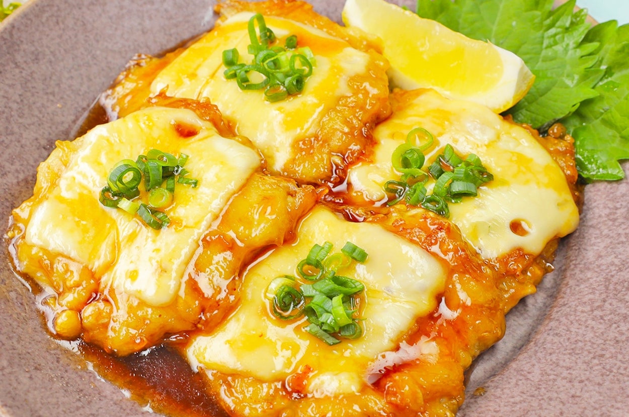 お皿に盛られたチーズがのった照り焼きチキンの一品。レモンと薬味が添えられている。