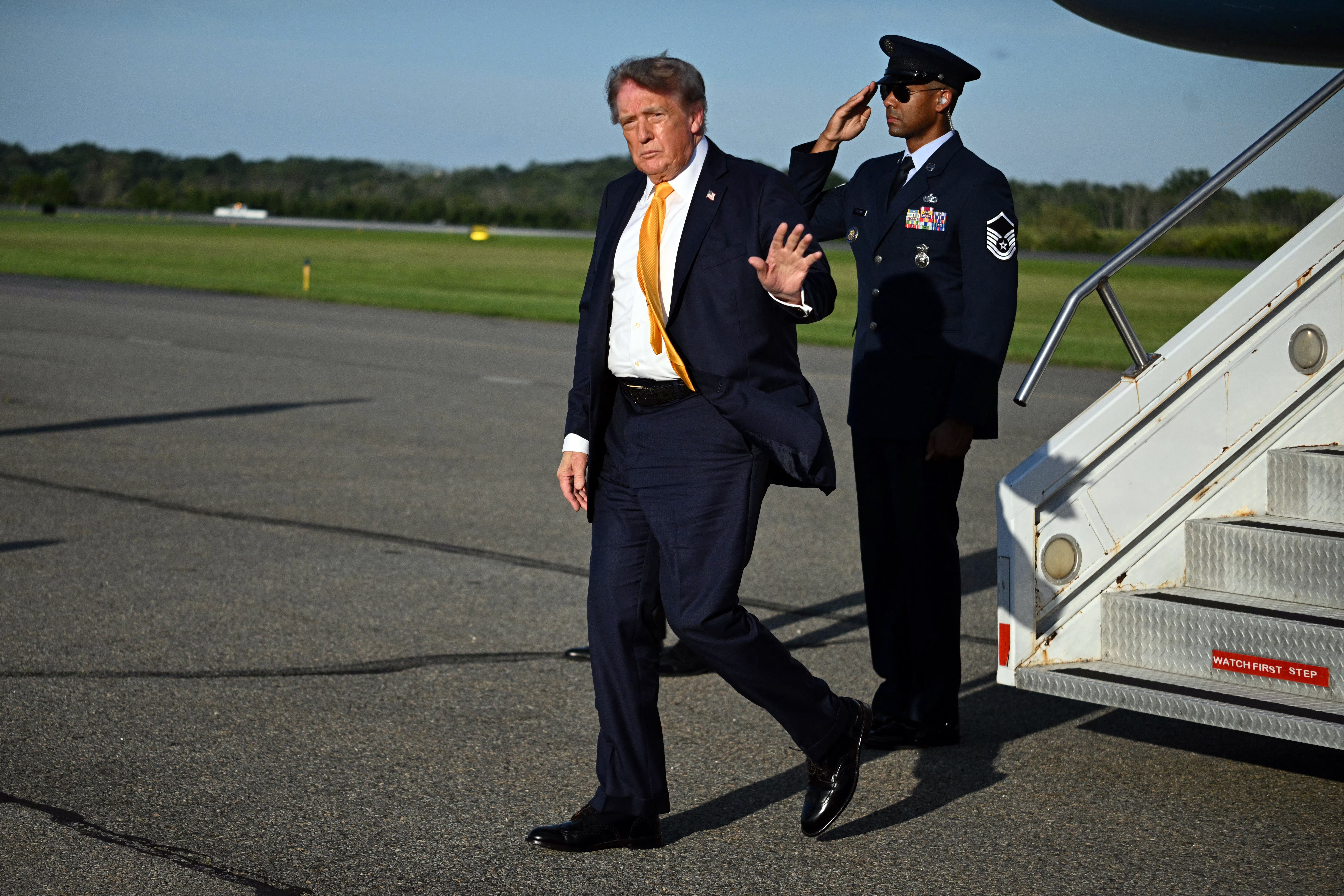 An unidentified man in a suit disembarks from a plane, waving while a uniformed individual salutes in the background on the tarmac