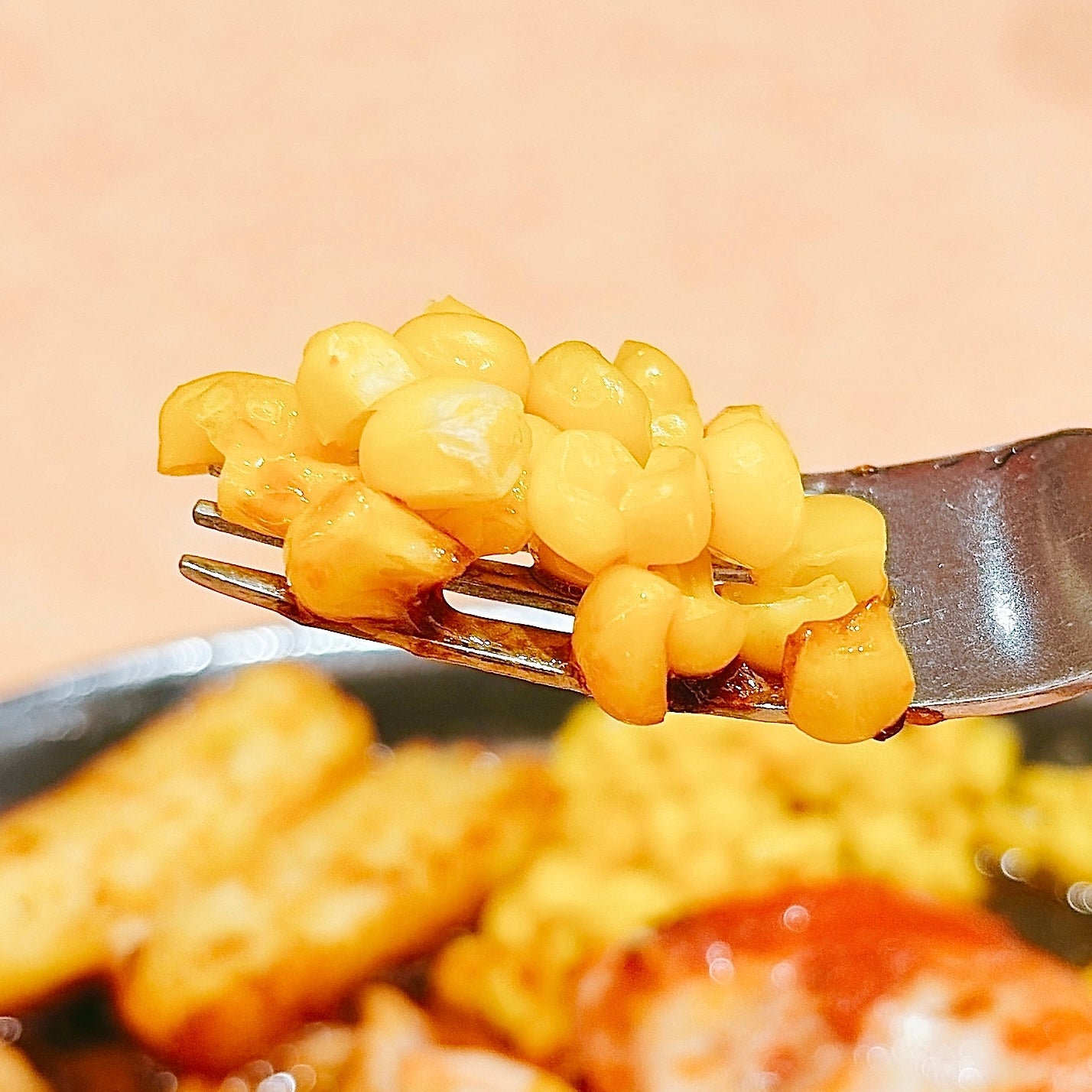 サイゼリヤ「柔らかチキンのチーズ焼き」フォークに盛られたコーンの粒。背景には他の食べ物がぼんやりと見える。