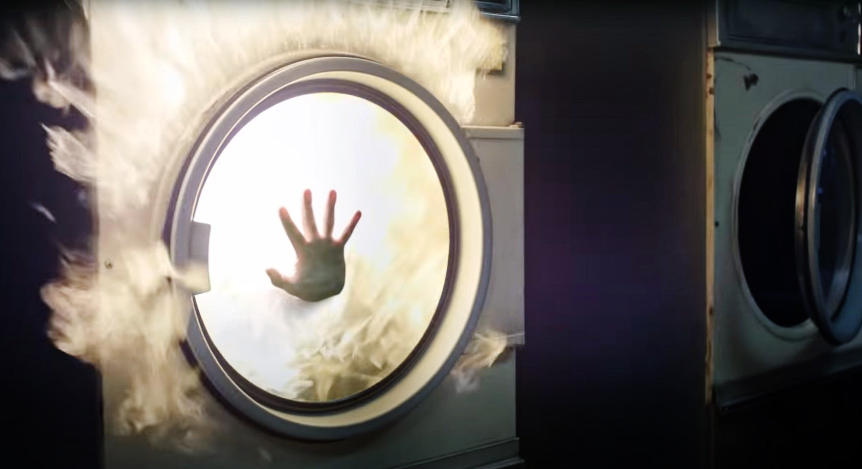 A hand presses against the window of a flaming dryer, illustrating a dramatic or hazardous situation in a laundry setting