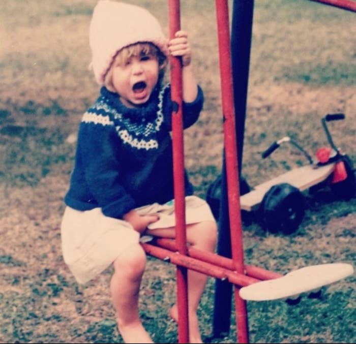 Child sitting on a red tricycle, wearing a cozy sweater and a knit hat, open-mouthed as if laughing or shouting