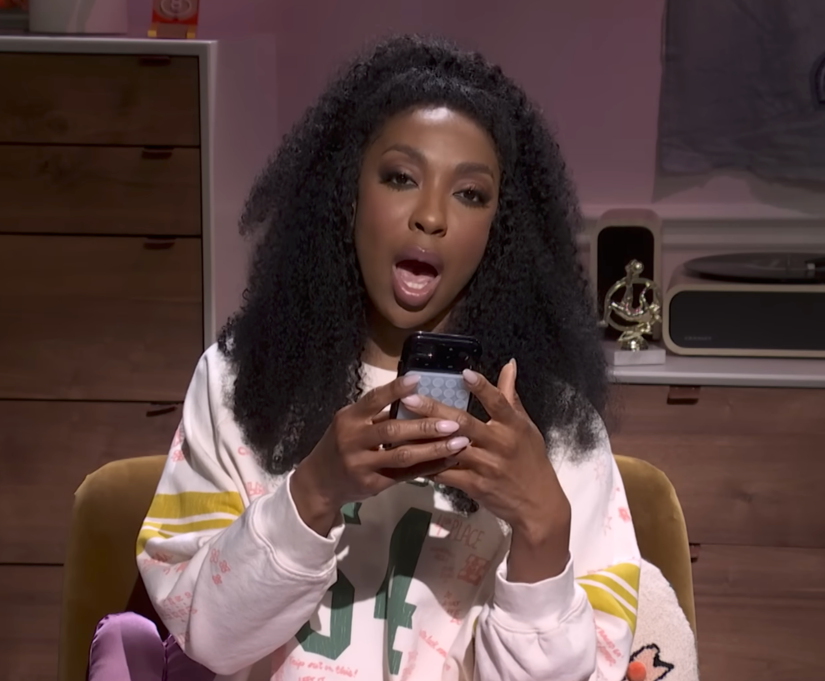 Person with long curly hair, wearing a casual sweatshirt, holding a phone while speaking. They appear to be in a home setting