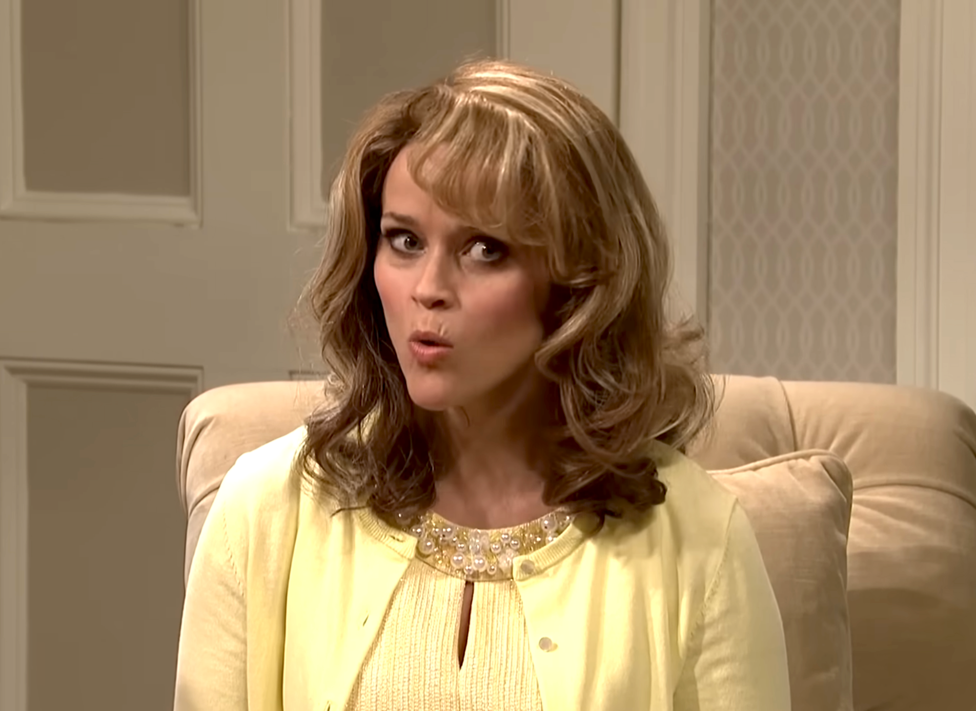 Person with medium-length hair, wearing a light-colored cardigan and top, sitting in a cushioned chair, making a surprised facial expression