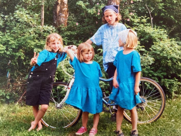 Four children pose playfully by a bicycle in a grassy area. Two girls in matching dresses and another in overalls smile and make peace signs