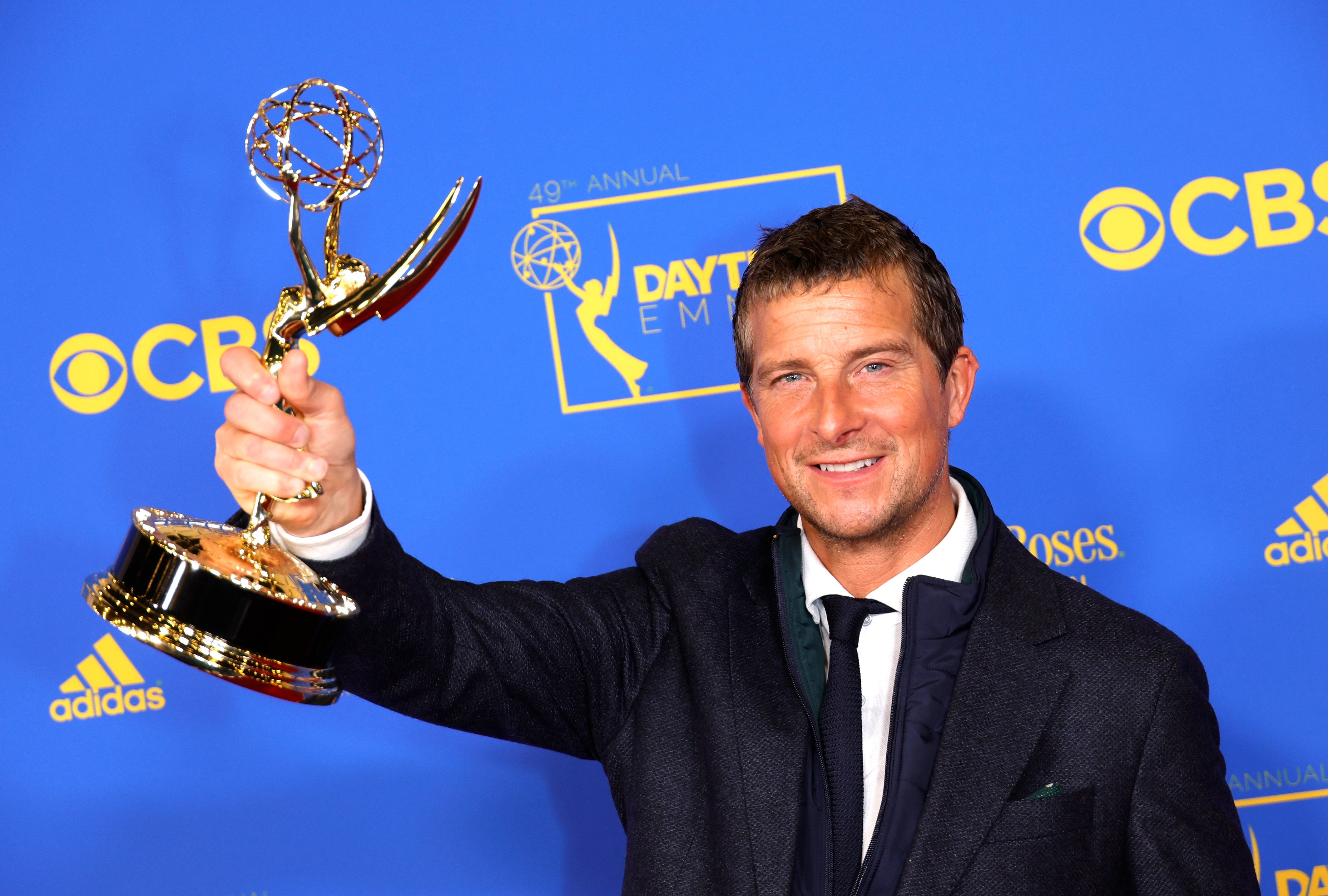 En person i dress holder en Emmy-pris, smilende, mot et blått bakteppe med Emmy-logoer, noe som tyder på en feiring ved en prisbegivenhet