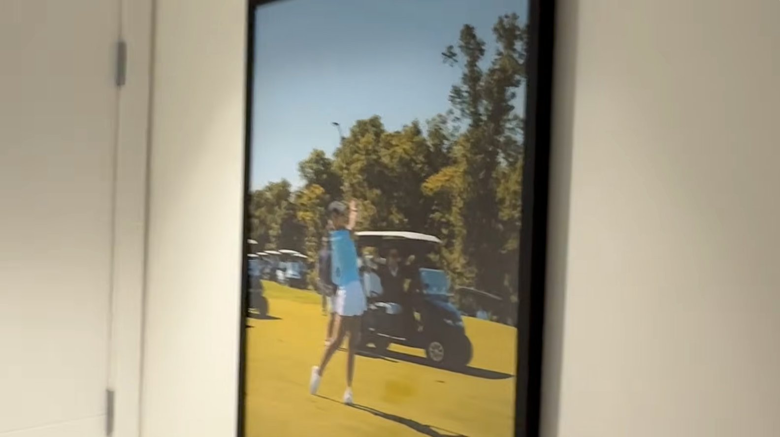 Et innrammet bilde på en vegg viser en person som spiller golf med golfbiler og trær i bakgrunnen