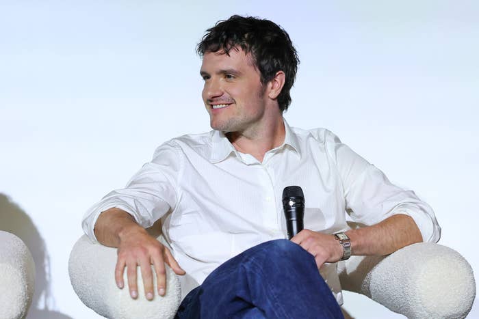 A person in a casual white shirt and jeans sits on a chair, holding a microphone, smiling at an event