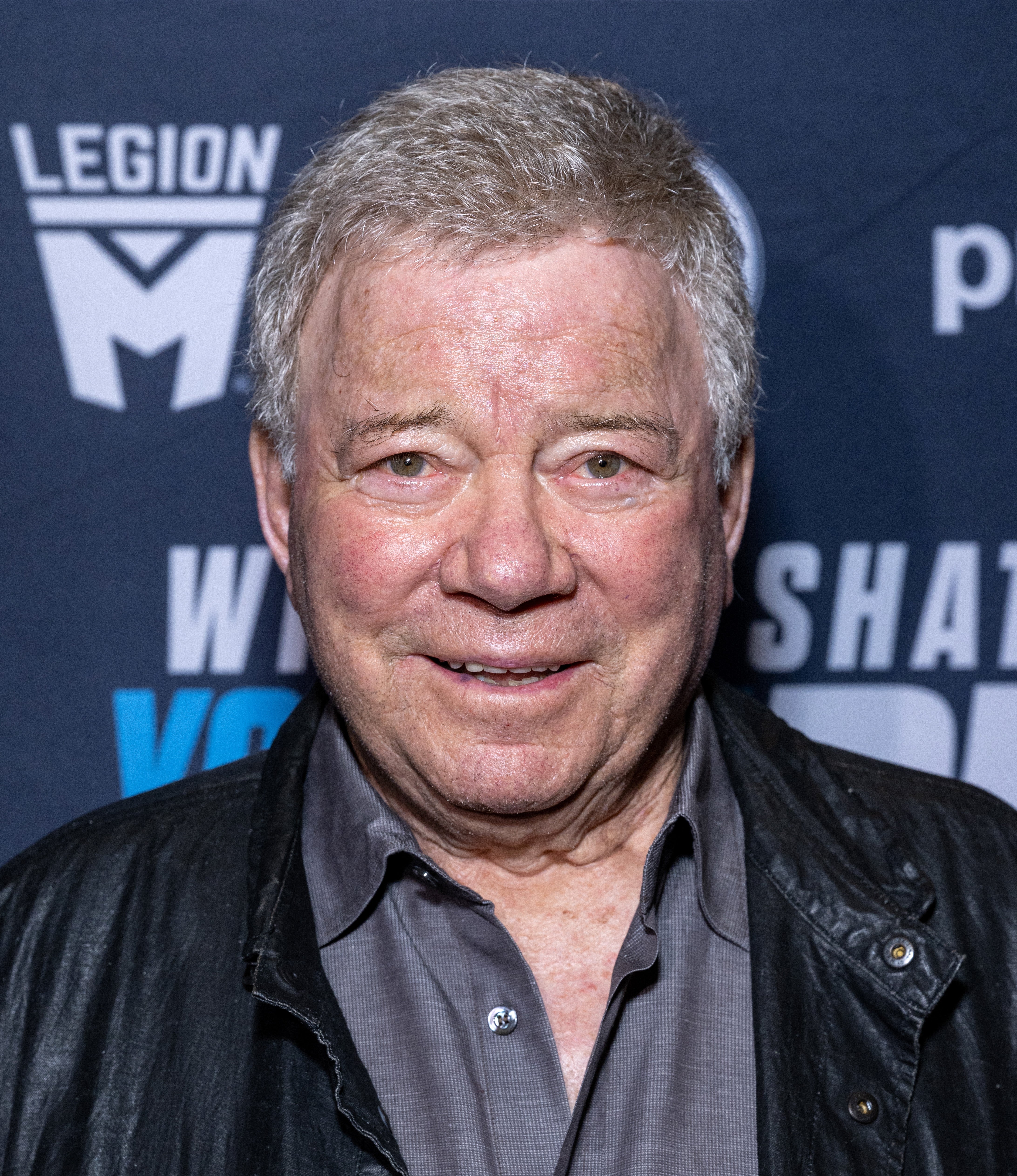 Person smiling at an event, wearing a leather jacket over a button-up shirt, standing in front of a backdrop with logos
