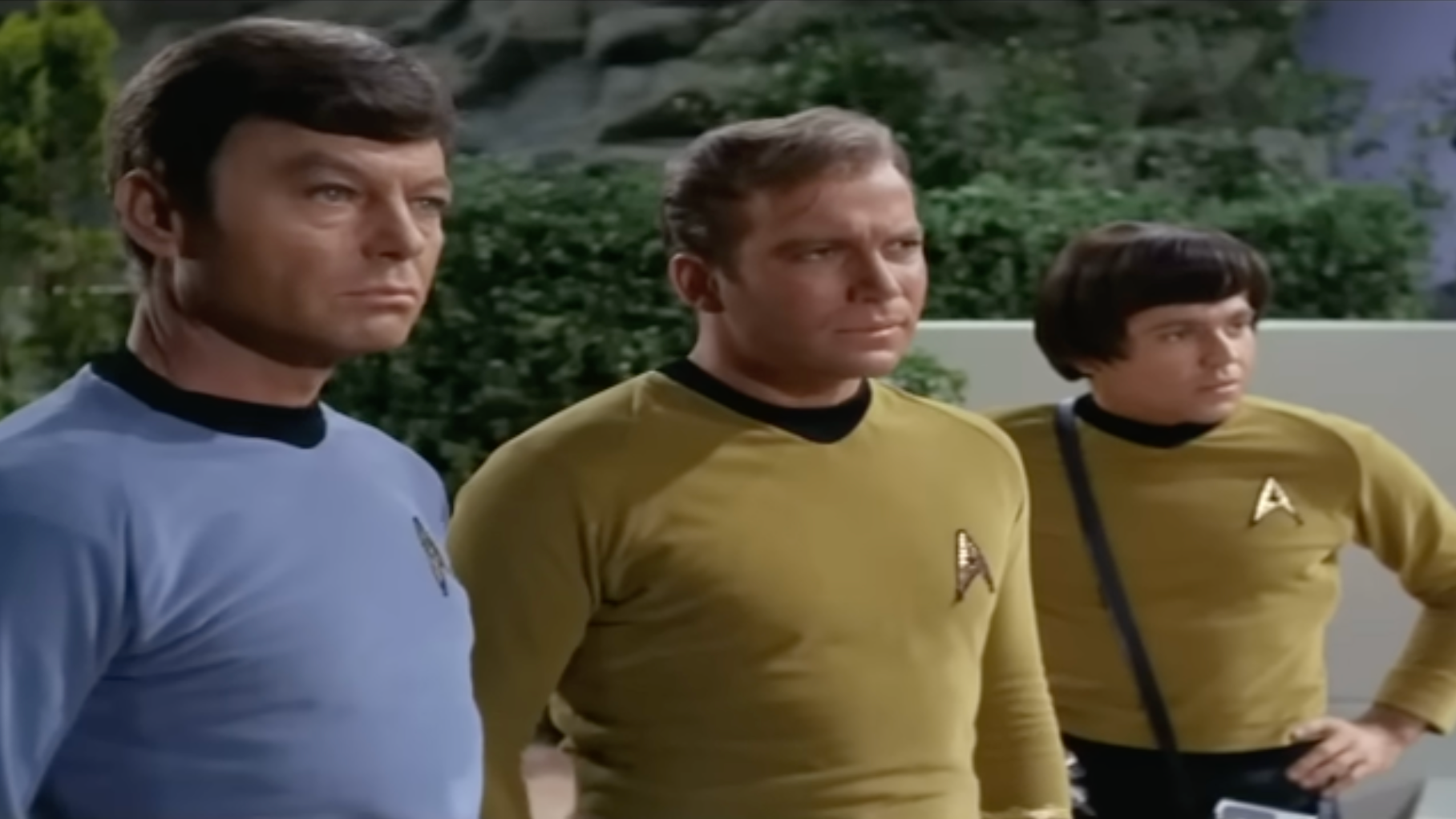 Three people in futuristic uniforms stand attentively, two in gold and one in blue, appearing focused and serious in an outdoor setting