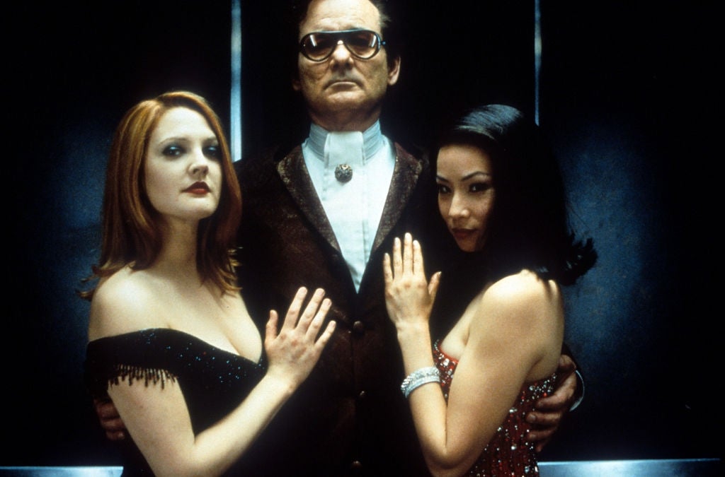 Two women in elegant dresses stand with a man in a suit. They pose confidently with their hands on his chest, evoking a classic Hollywood vibe