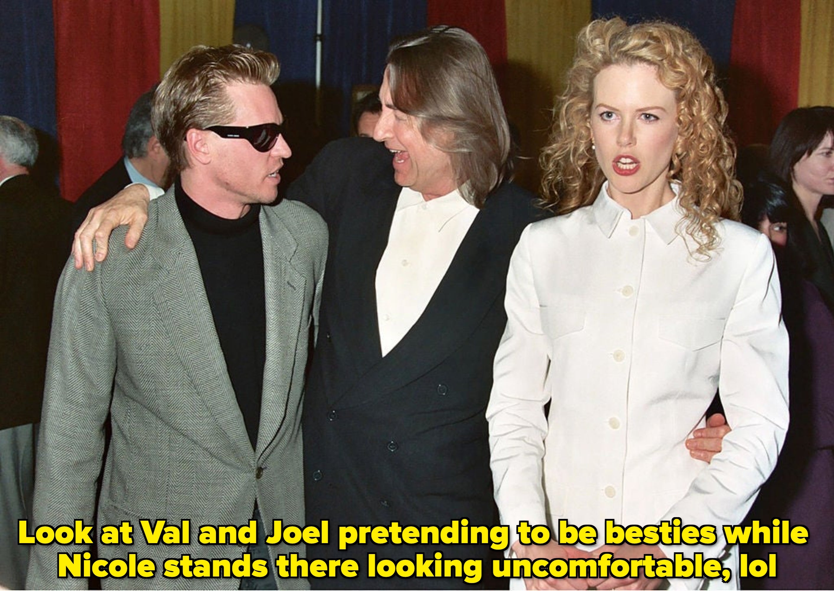 Three people are socializing at an event. One wears sunglasses and a suit, another wears a suit, and the third wears a white outfit with curly hair