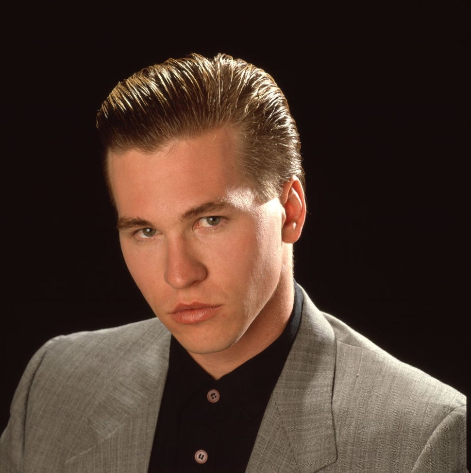 A person in a suit jacket and black shirt gazes forward. Slicked-back hair gives a polished appearance