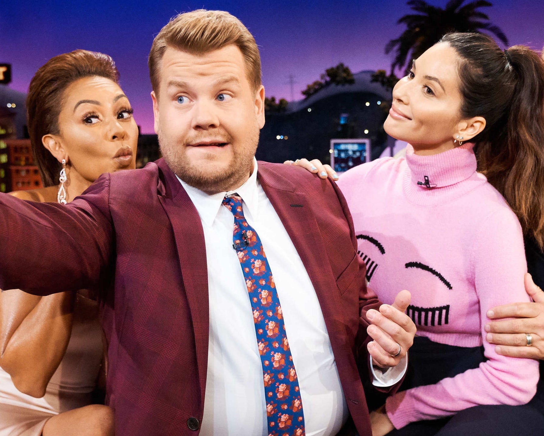 People pose for a group selfie on a talk show set. One person wears a suit, another a pink sweater with a face design, and others wear casual outfits