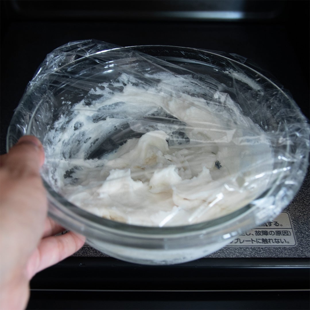 電子レンジに入れられたラップで覆ったガラスボウルに白い生地が入っている。