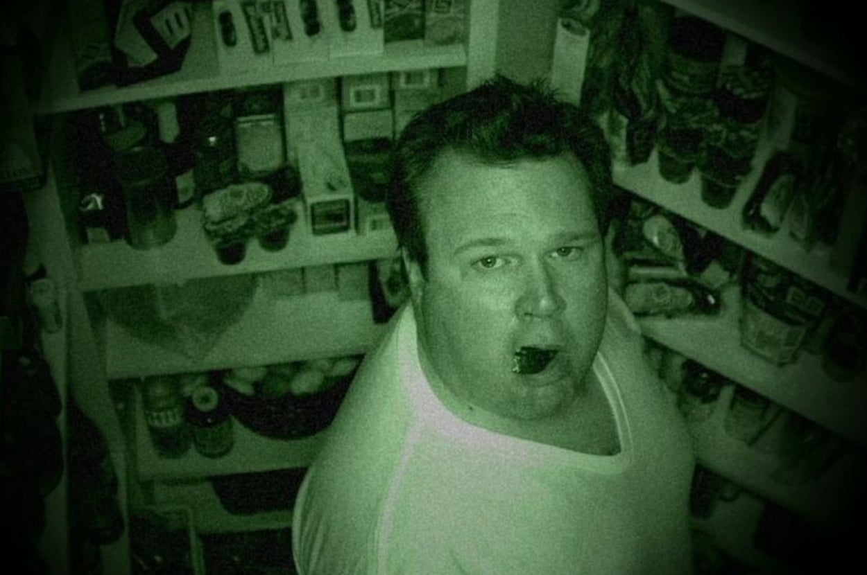 A man looks surprised while caught eating in a pantry at night, surrounded by various food items