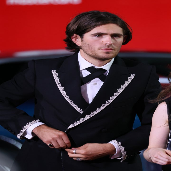 Person in a formal suit with intricate lace trim, standing on a red carpet