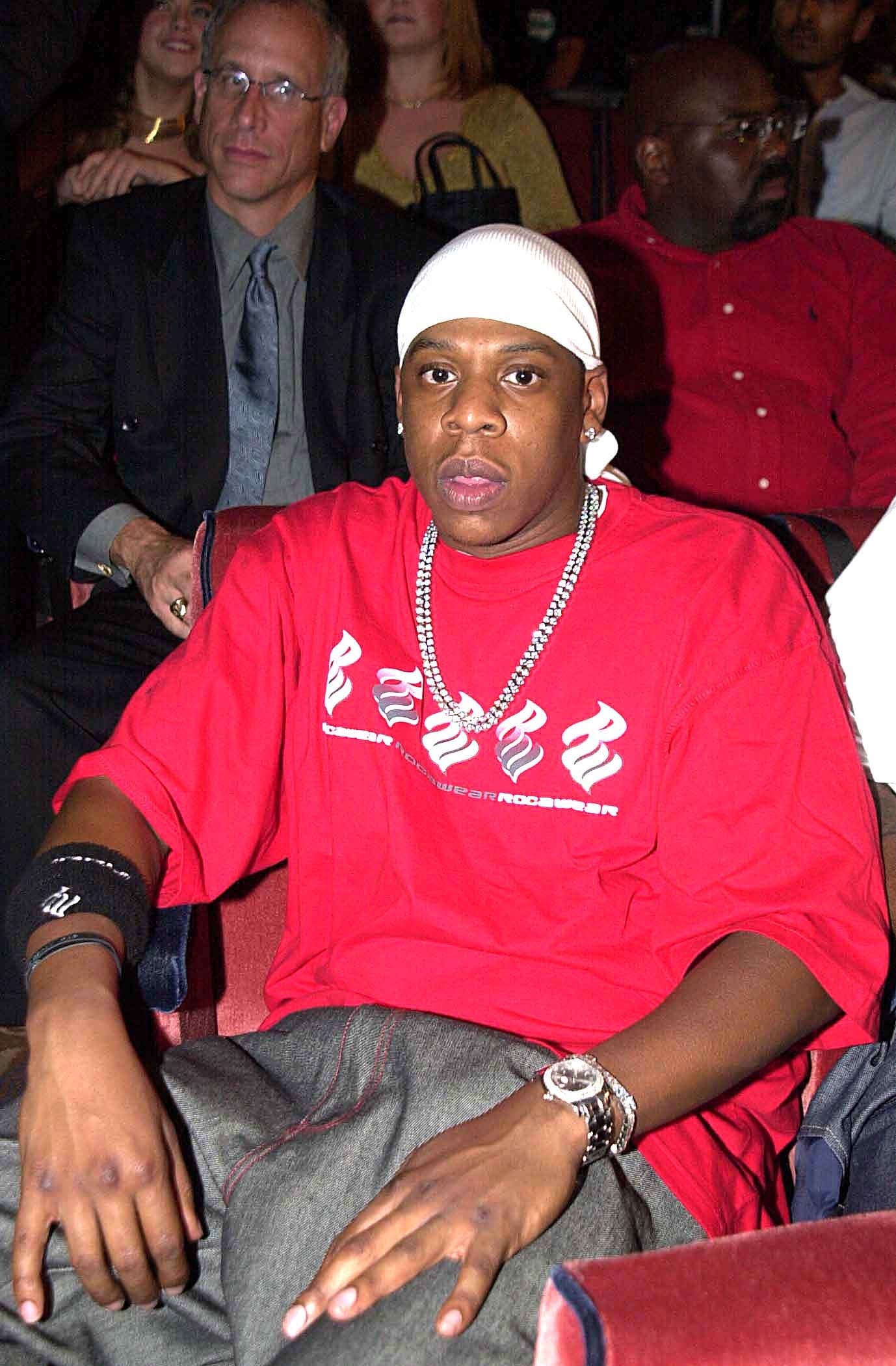 Person in a durag and chain necklace wearing a red oversized shirt with logos, sitting in an audience