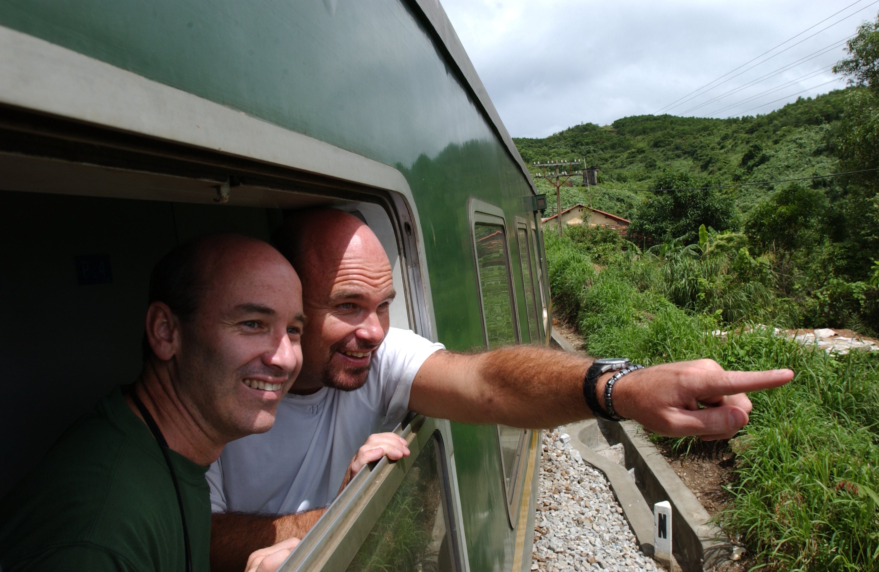 Two people smiling and looking out the window of a moving train, enjoying the scenic green landscape outside