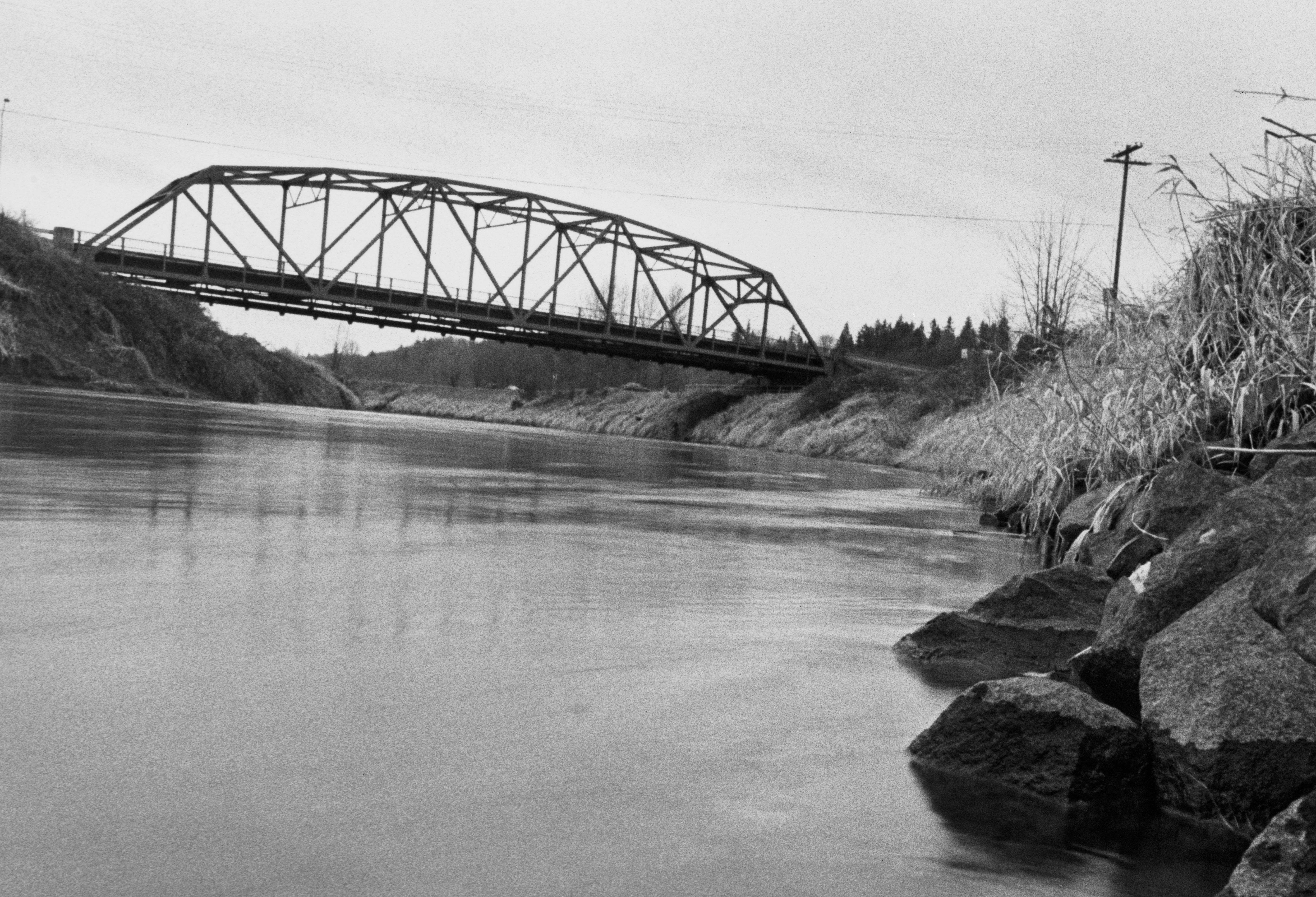 Black and white photo of a metal bridge over a calm river, with grassy banks and rocks in the foreground