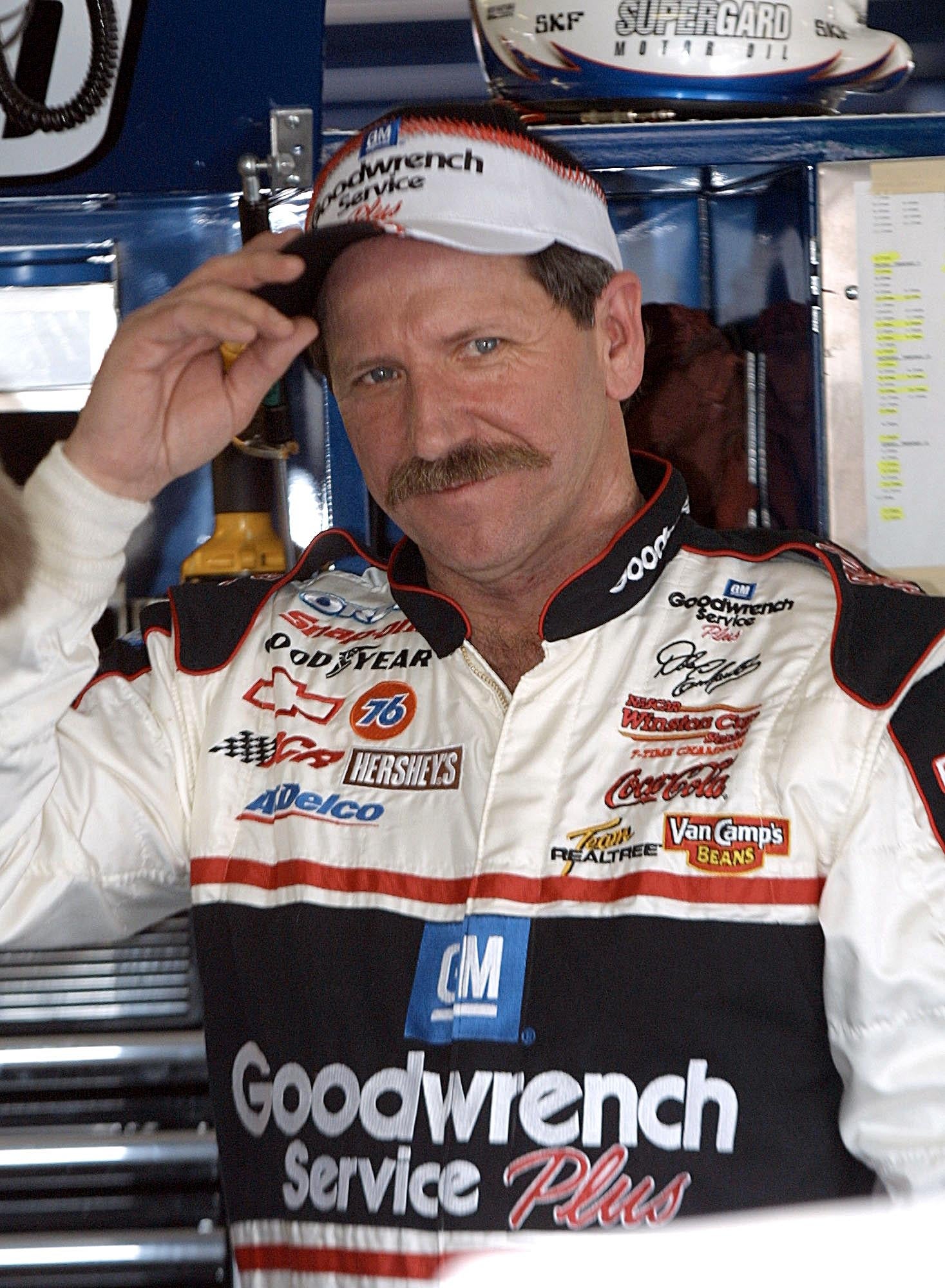 Person in a racing suit with sponsor logos, wearing a cap, slightly smiling and tipping the cap