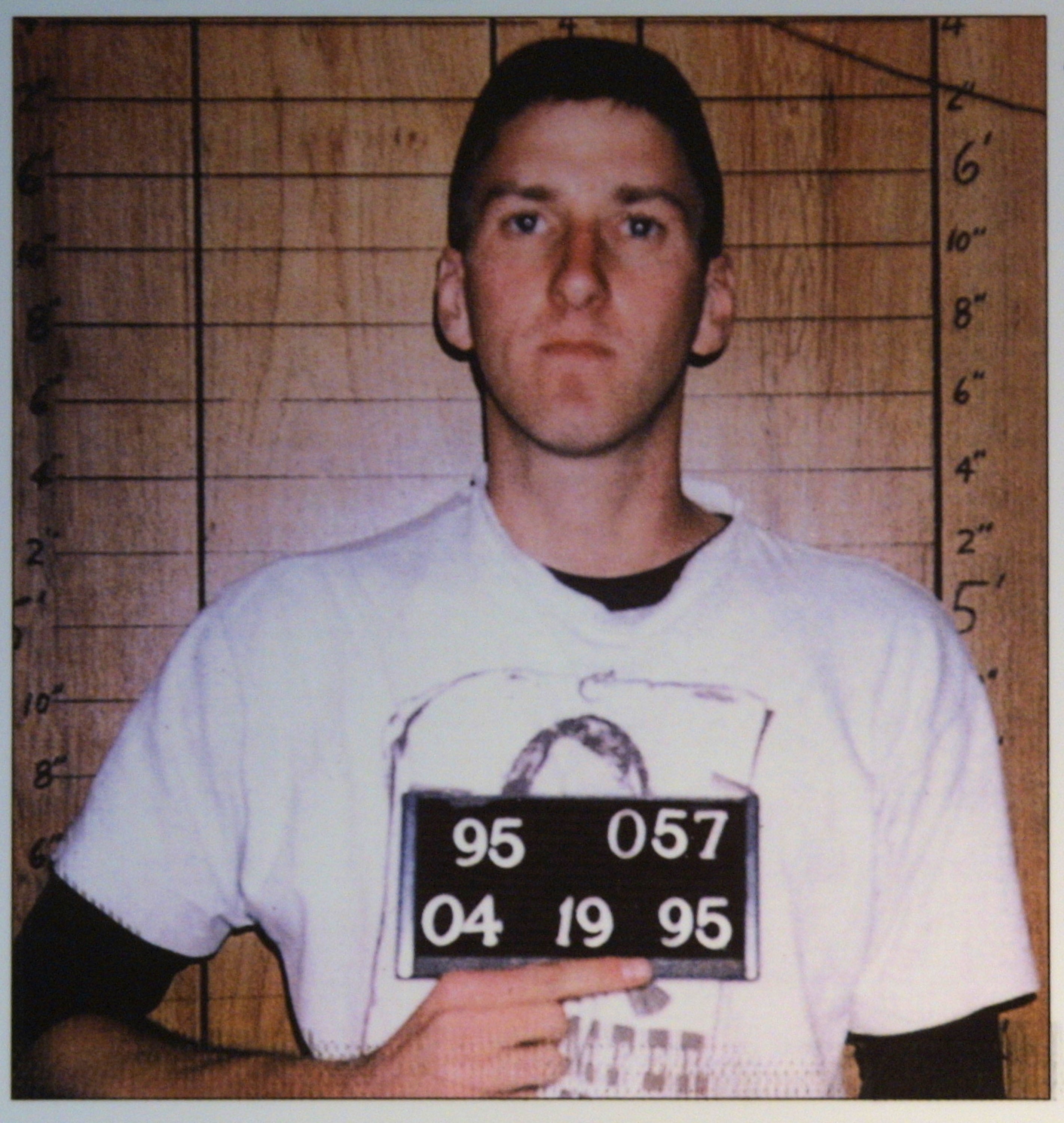 A person holding a sign with their date and identification numbers for a mugshot, standing against a height measurement backdrop