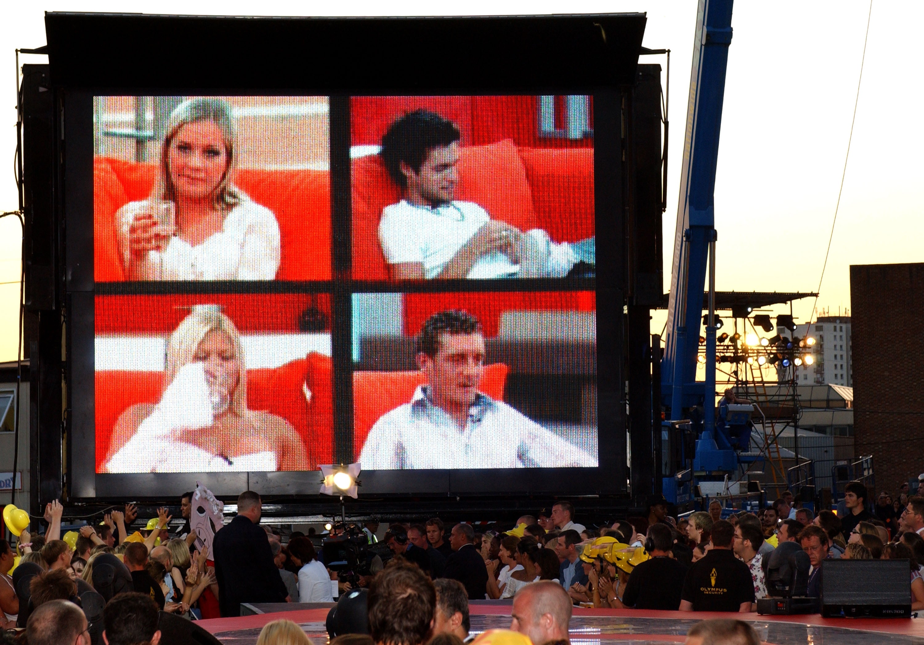 Large outdoor screen showing four people seated indoors, with a crowd gathered below watching the display
