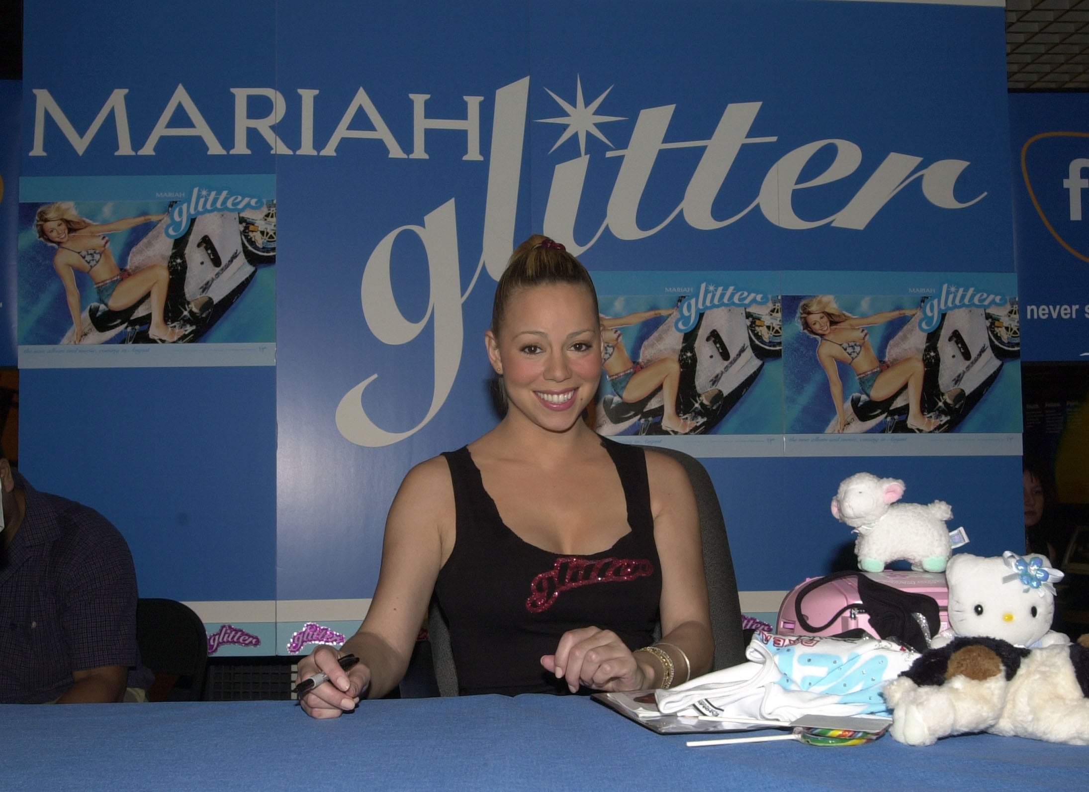 A person sits at a table with plush toys and promotional materials, in front of a "Glitter" poster backdrop