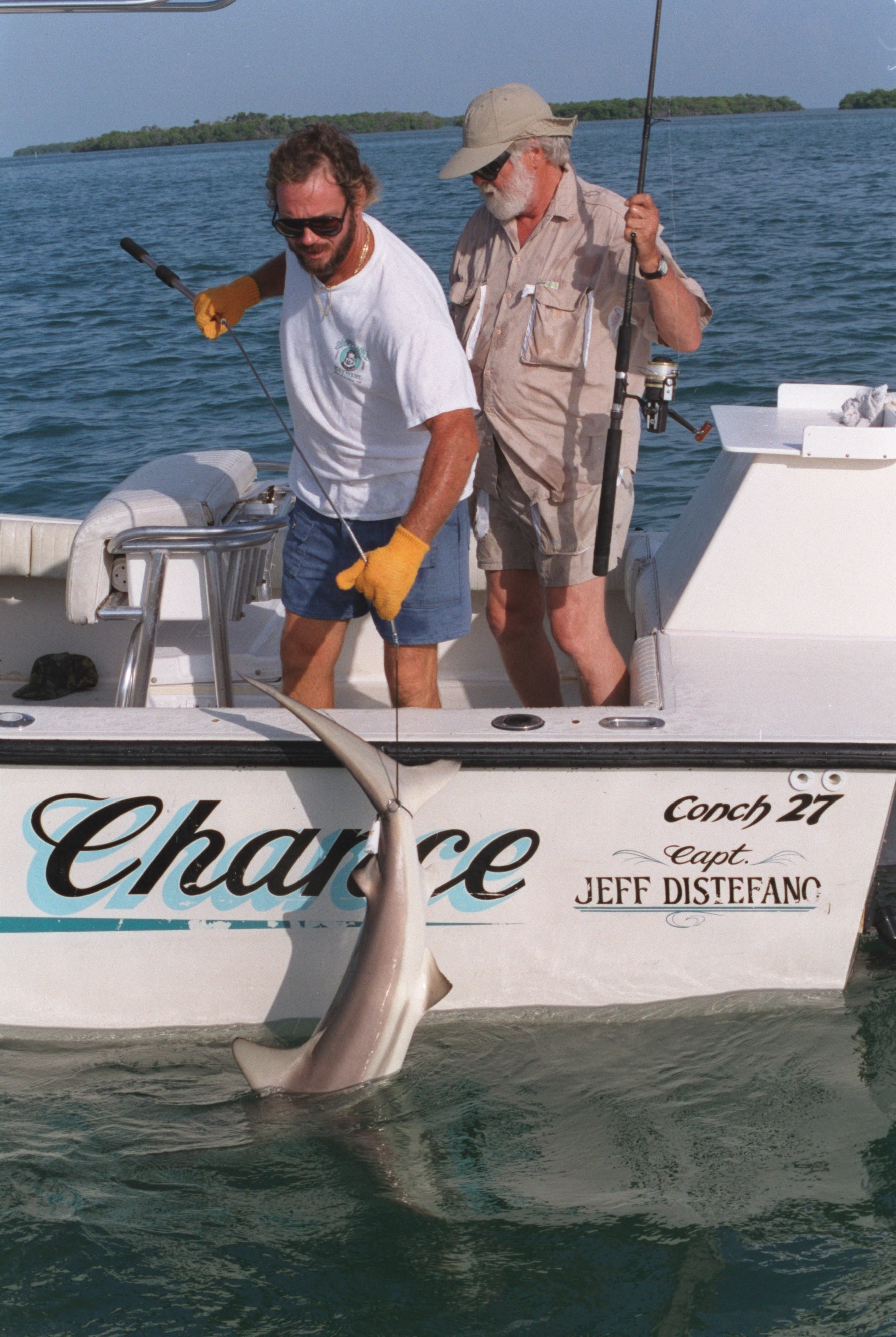 Two men on a boat catch a small shark. One wears casual shorts and a T-shirt, the other in a sun hat and fishing gear, both engaged in fishing activity